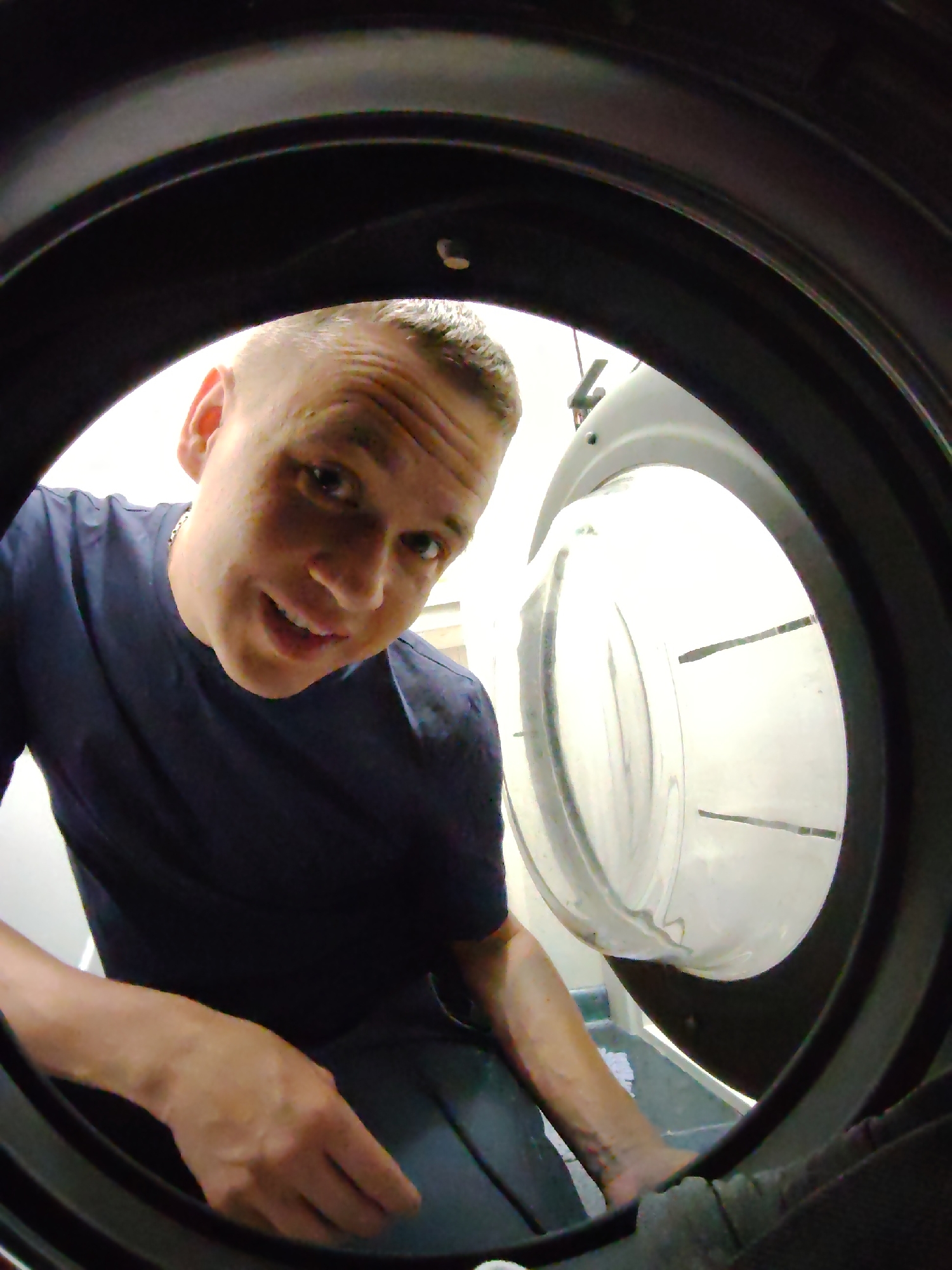 A man looks into a washing machine, smiling slightly. He is wearing a dark blue t-shirt and a silver chain. His hair is cut short. His left hand reaches into the washing machine drum. The washing machine door is open. The interior of the washing machine drum is visible. The scene is in Chaikovsky, Russia.