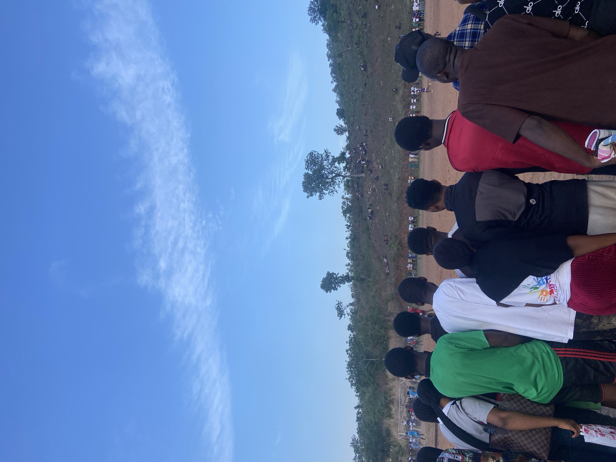The image shows a group of people standing in a line, looking out over a field in Lokoja, Nigeria. A crowd of people, including children, have gathered, possibly to watch a soccer match. There is a hill and some trees in the background, and the sky is clear and blue.
