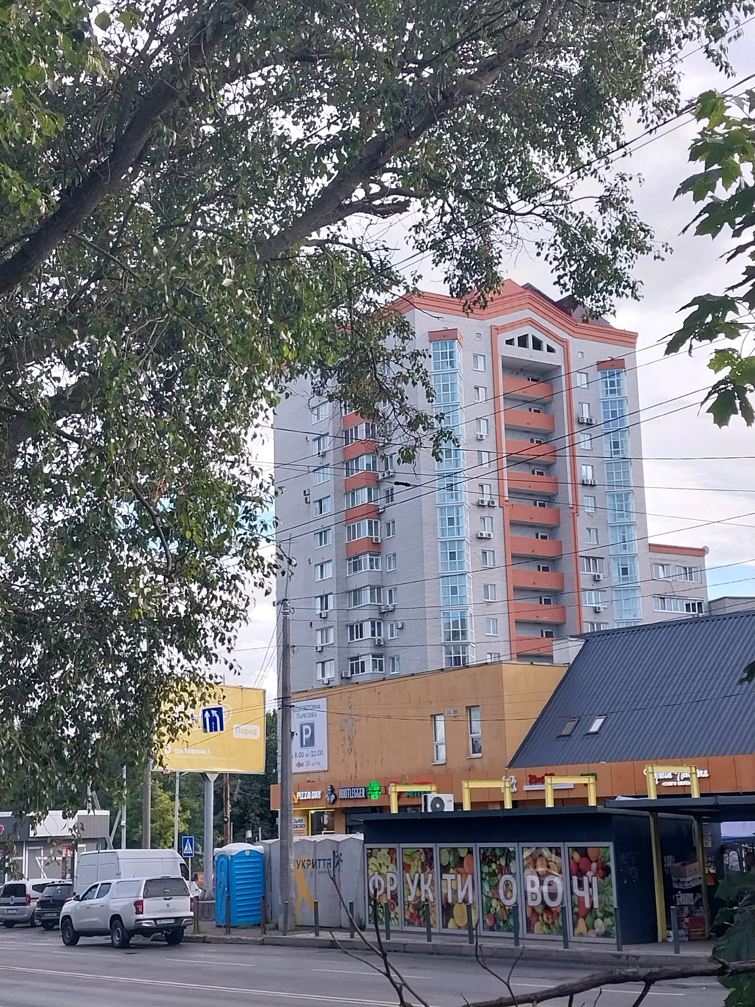 The image depicts a street scene in Dnipro, Ukraine, focusing on a modern, light-colored apartment building with orange accents and balconies. The building is tall, with numerous windows and air conditioning units visible. In the foreground, a street with parked cars, including a white pickup truck and smaller vehicles, runs alongside a sidewalk. There are several portable toilets on the sidewalk, a yellow billboard with blue signage, and a row of shops with various signs, one of which displays images of fruits and vegetables. Overhead, tree branches with green leaves partially obscure the view of the sky, which appears overcast. Power lines are also visible, crisscrossing the scene. The overall impression is a typical urban environment, with a mix of residential and commercial structures.