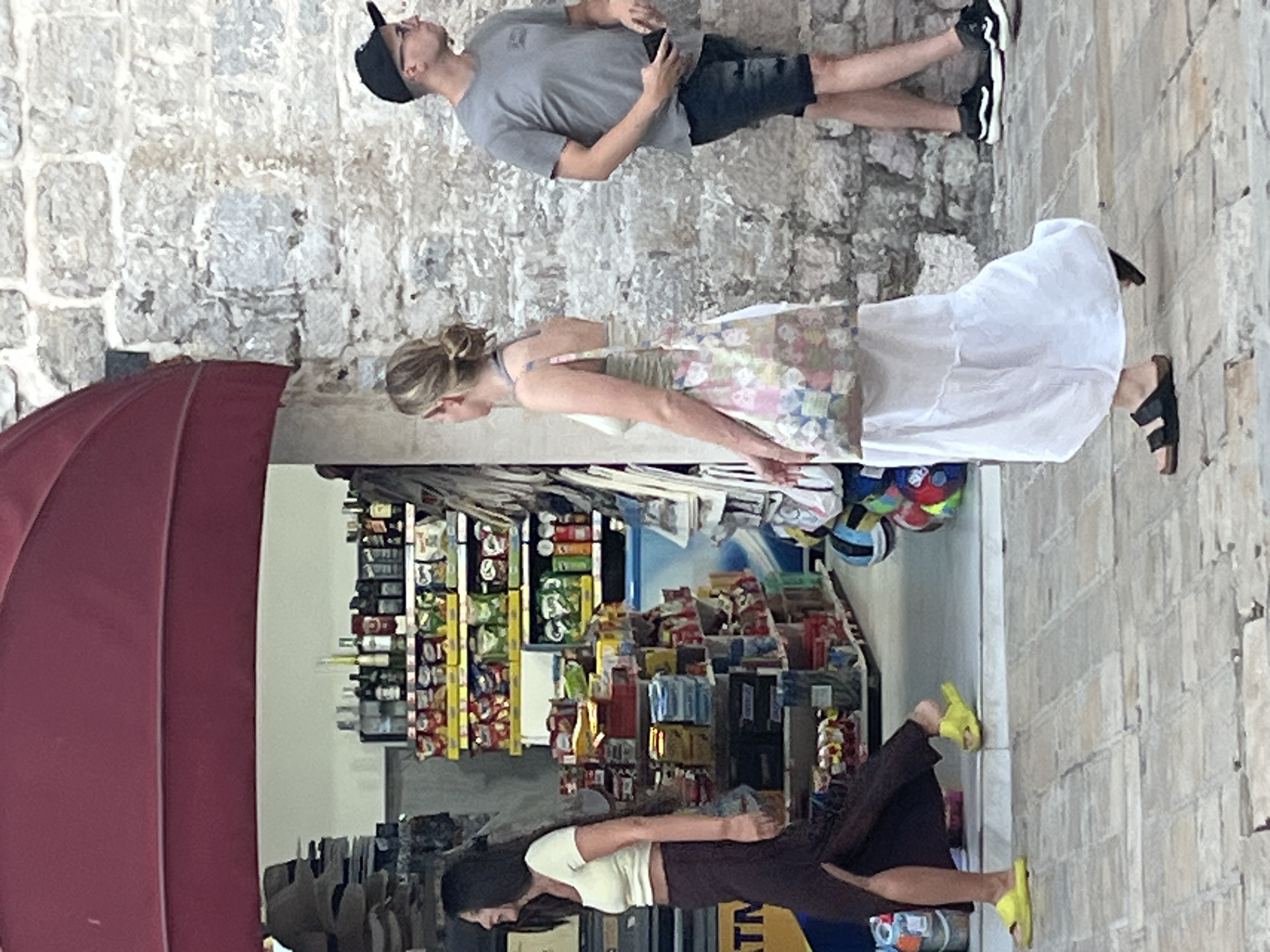 A group of people are walking along a paved street in Budva, Montenegro. On the left, a woman with long dark hair wearing a white crop top and a dark, high-slit skirt walks past a storefront. In the center, a woman with blonde hair in a bun, wearing a white tank top and a long white skirt, carries a colorful tote bag. To the right, a man wearing a grey t-shirt, black shorts, and a baseball cap walks by. The storefront behind the woman on the left is stocked with various goods. The background features a stone wall and a red awning.