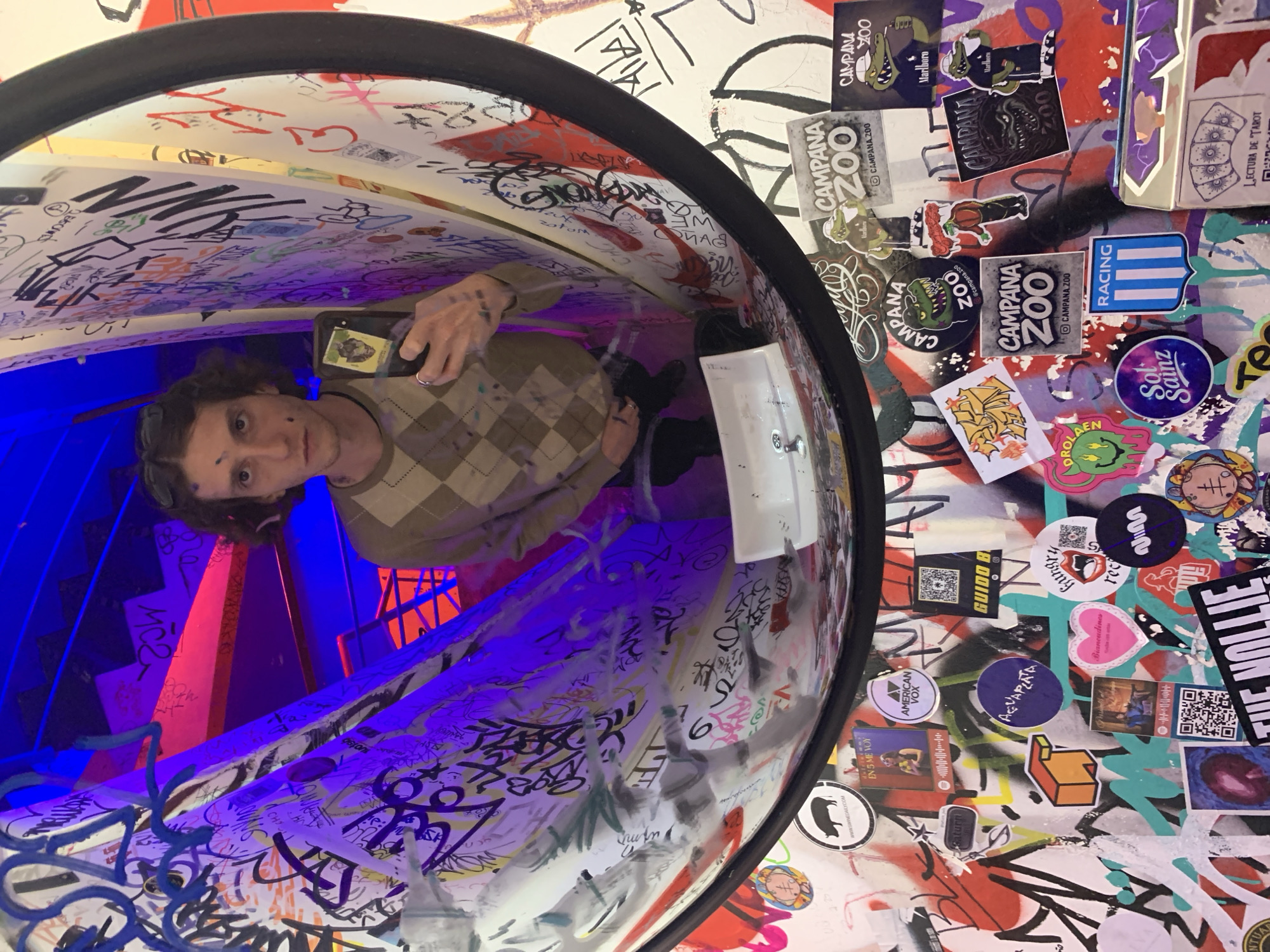 Here is a forensic description of the image:
The image is an eye-level, medium shot of a person taking a photo of themself in a periscope-like art installation. The subject, presumably male, is partially visible from the torso up, facing the camera. He has brown hair and is wearing an argyle sweater. He is holding a mobile phone in his right hand.
The background of the installation is covered in graffiti, stickers, and tags. A red and blue light installation is visible within the periscope. Stickers with the words "CAMPANA ZOO" are noticeably present, along with other miscellaneous stickers. The installation itself is cylindrical and black around the outer rim.
The location is Buenos Aires, Argentina.