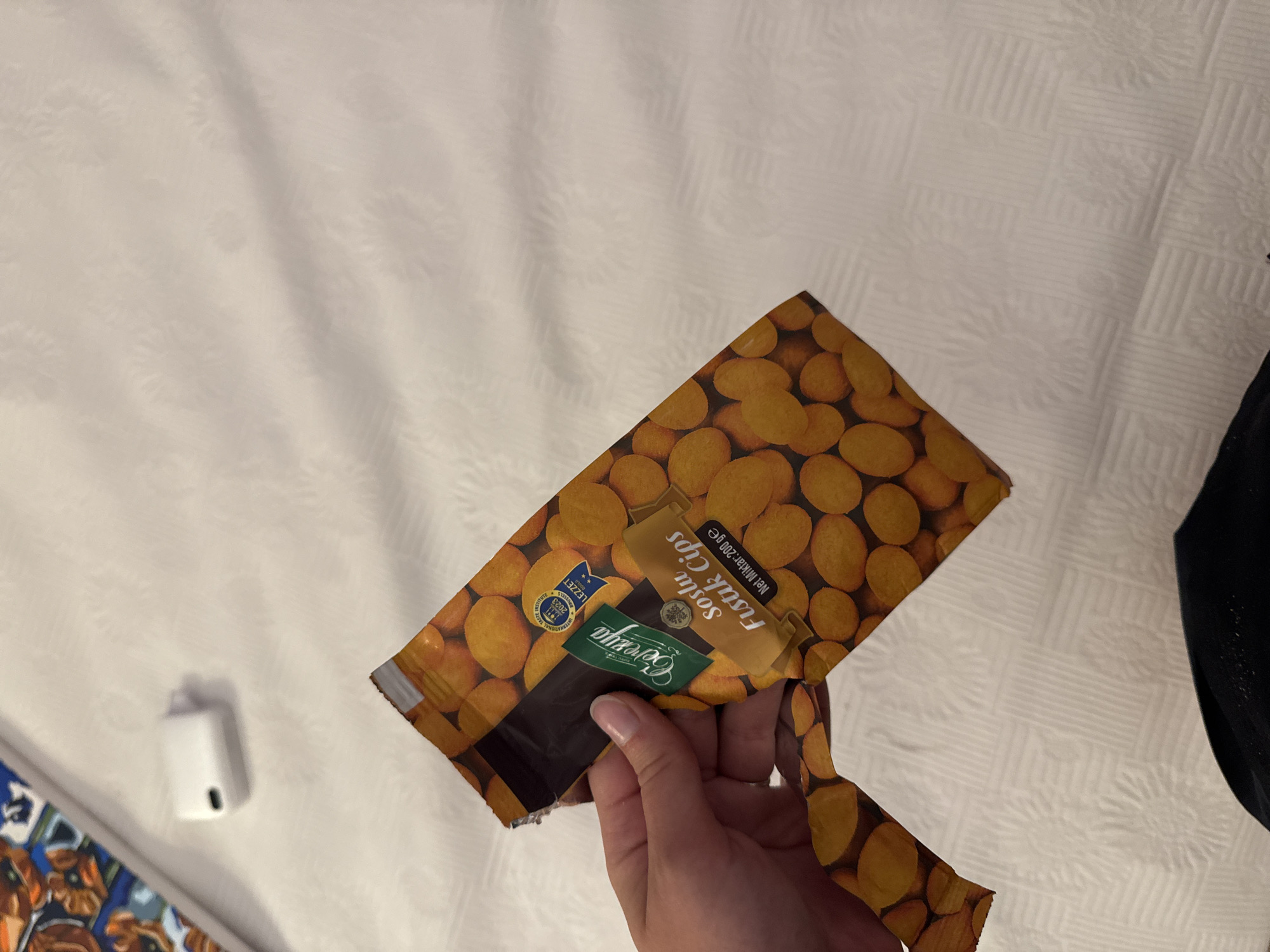 A hand holds a torn package of "Soslu Fıstık Cips" (Spiced Peanut Chips) manufactured by "Cerezya" from Turkey. The package, featuring an image of peanuts and Turkish text, is partially open. The scene is set on a white quilted surface, possibly a bed or sofa, with a blurred colorful fabric in the upper left corner. A white electronic device, resembling an earbud charging case, is visible in the upper portion of the image. The specific location within Aksu, Turkey, cannot be determined from the visual information provided.