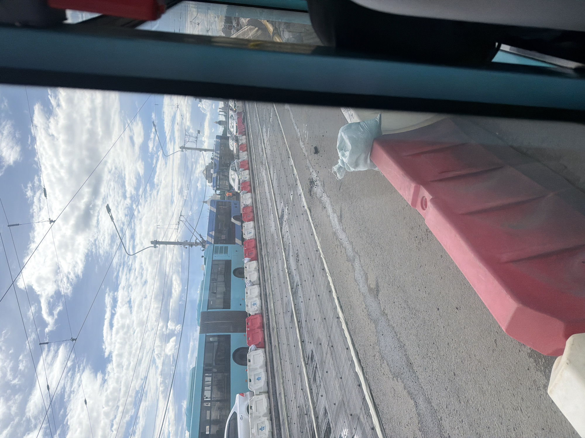 A view from inside a public transport vehicle, likely a tram or trolleybus, looking out at a street scene in Saint Petersburg, Russia. The foreground shows a red plastic barrier and a white bag filled with sand, suggesting ongoing construction or road work. Beyond the barrier, a wide road is visible with visible tram tracks embedded in the asphalt, which is wet in places. Several turquoise-colored public transport buses are lined up on the far side of the road. Other vehicles, including cars and trucks, are also present, some of which appear to be stationary in traffic. Overhead, a network of power lines and poles stretches across the sky, characteristic of Saint Petersburg's urban infrastructure. The sky is partly cloudy with a mix of blue sky and white clouds. In the distance, several buildings and church domes are visible against the horizon.