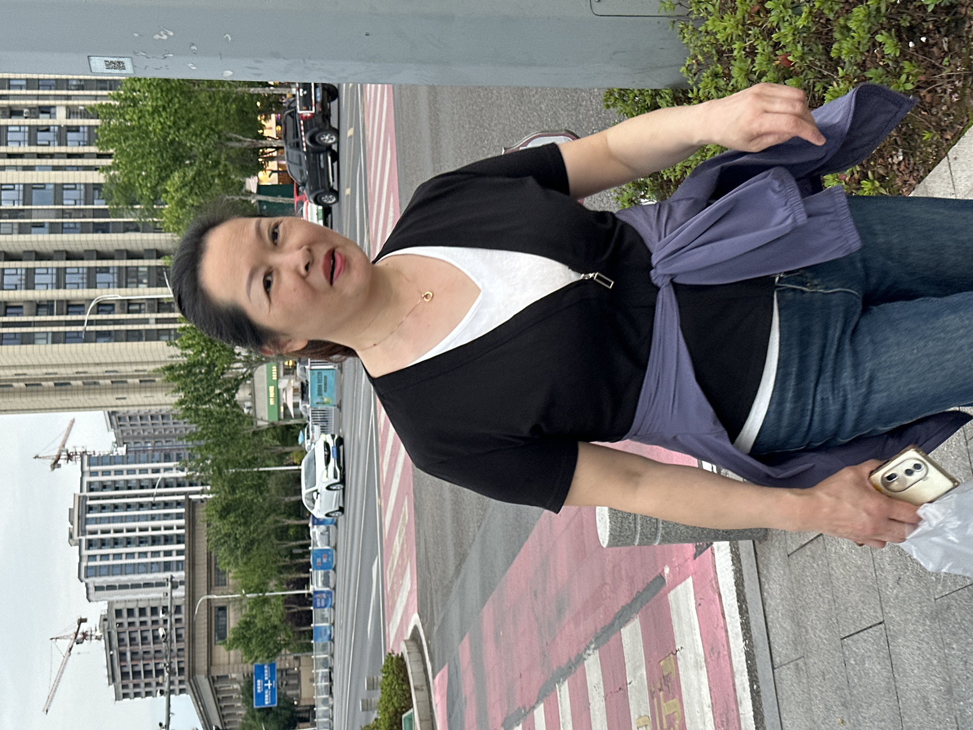 A woman stands on a sidewalk in Jiangjin, China, holding a smartphone in her right hand and facing the camera. She wears a black cardigan over a white top, blue jeans, and a lilac-colored wrap around her shoulders. A street with a crosswalk is behind her. Buildings, trees, and cranes are visible in the background, indicating an urban environment.