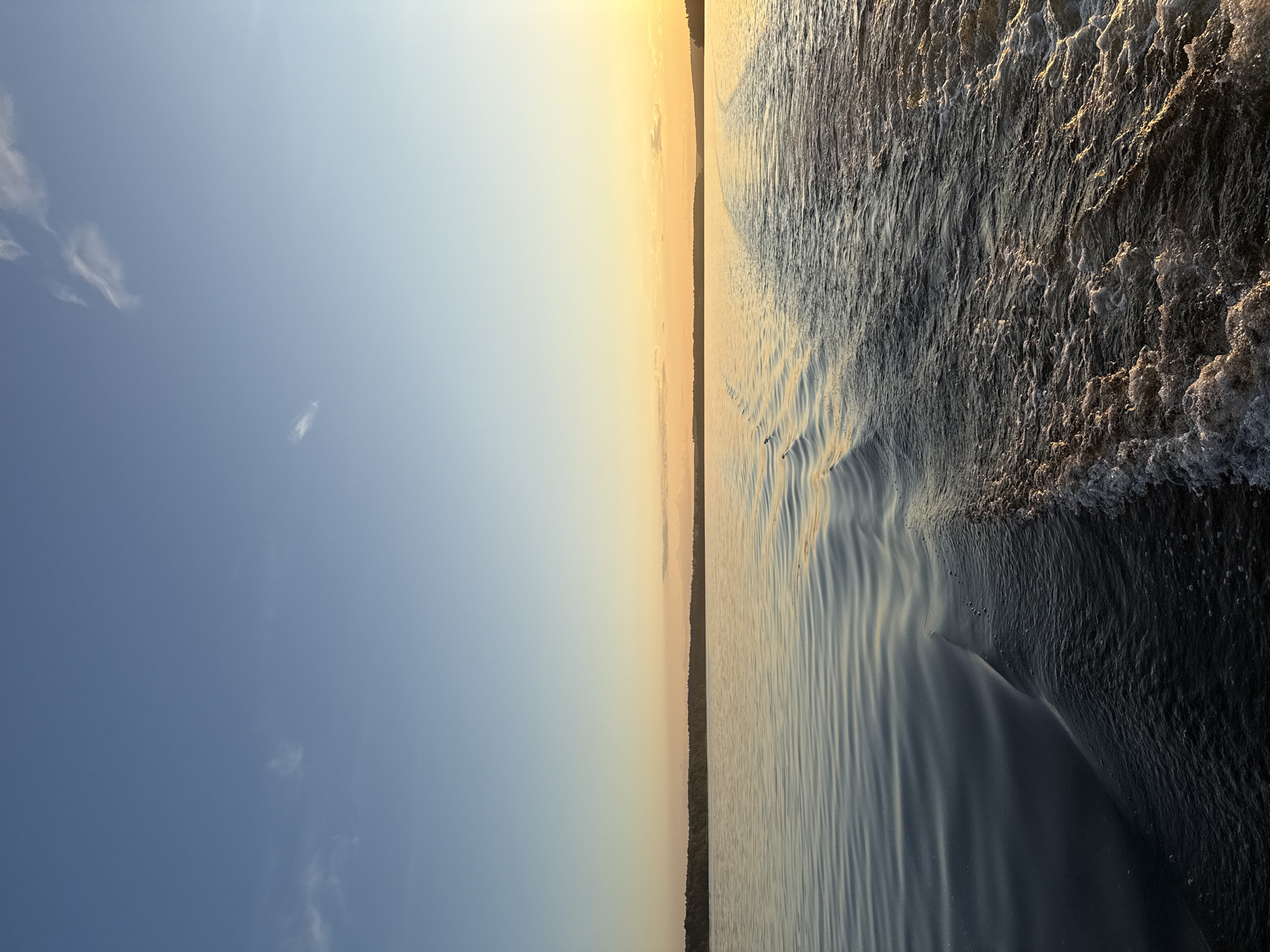 This image depicts a natural landscape at either sunrise or sunset over a large body of water, located within the vicinity of Votkinsk, Russia. The scene is dominated by a clear sky, a wide expanse of water, and a distant forested shoreline.
The sky occupies the upper portion, displaying a gradient of color. It transitions from a light, clear blue in the upper left to a warmer, more yellowish-orange hue towards the right, indicative of a low sun. Sparse, thin cirrus clouds are visible in the upper half of the sky.
A dark, low-lying band of dense forest stretches across the horizon, separating the sky from the water. This landmass appears consistent and unbroken along the visible horizon.
The water fills the lower two-thirds of the image. On the left side, the water's surface is relatively calm, characterized by subtle, undulating ripples that reflect the soft blues and yellows of the sky. On the right side, a distinct, turbulent wake of white foam and disturbed water extends from the foreground towards the distant horizon, suggesting the recent passage of a boat or the viewpoint from a moving vessel. This wake is illuminated by the golden light source on the right. No people, specific man-made structures (other than the boat wake), or discernible wildlife are present in the frame. The lighting conditions suggest the "golden hour" of early morning or late evening. If this scene is in Votkinsk, Russia, it likely portrays the Votkinsk Reservoir.