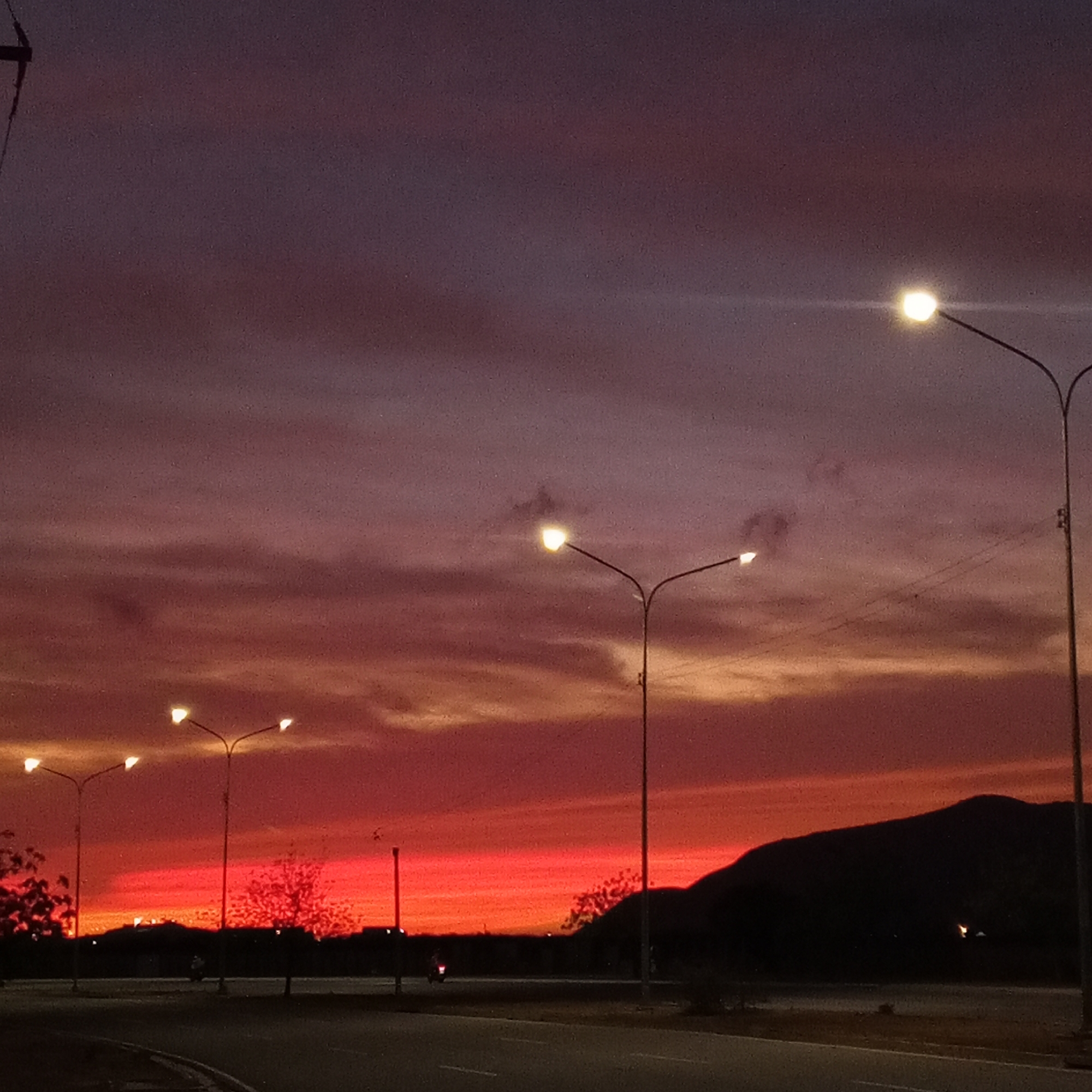 The image depicts a cityscape at sunset. The sky is a gradient of colors, ranging from deep red at the horizon to purplish-grey higher up. There are several streetlights illuminating the road. A dark silhouette of a hill or mountain is visible in the background. There are trees on the road that are lit with the sunset colors. A person is on a motorcycle on the road. The location is the city of Unknown, Unknown.