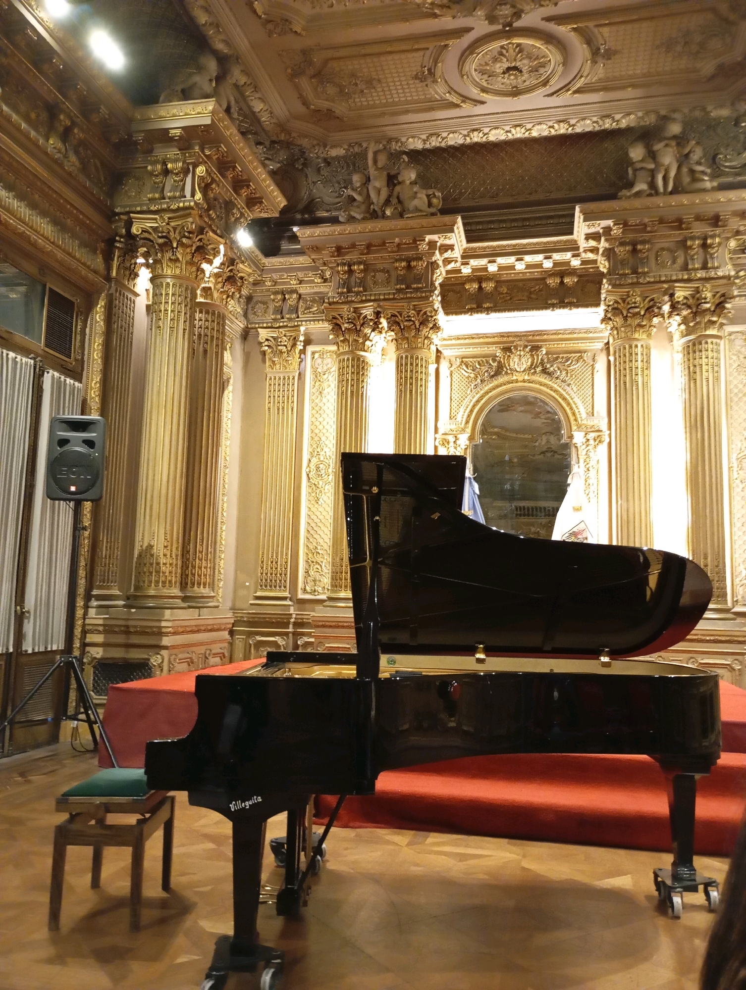 The image depicts a grand piano on a red carpeted stage within a richly decorated hall. The hall features ornate gold trim on the ceiling and walls, with elaborate columns and decorative figures. To the left of the stage, a black speaker on a tripod is visible, along with vertical striped curtains. A small, green-topped piano bench sits in front of the piano. The piano itself is black and glossy, with the brand name "Villeguita" inscribed on its side. The floor is made of polished wood. The scene appears to be set up for a performance or event in an opulent venue, likely the Salón Dorado of the Teatro Colón in Buenos Aires, Argentina, given the distinctive architectural style and the presence of the "Villeguita" piano, a brand historically associated with the theater. No people are clearly visible in the frame, though a sliver of a person's shoulder and hair is visible on the far right edge of the image.