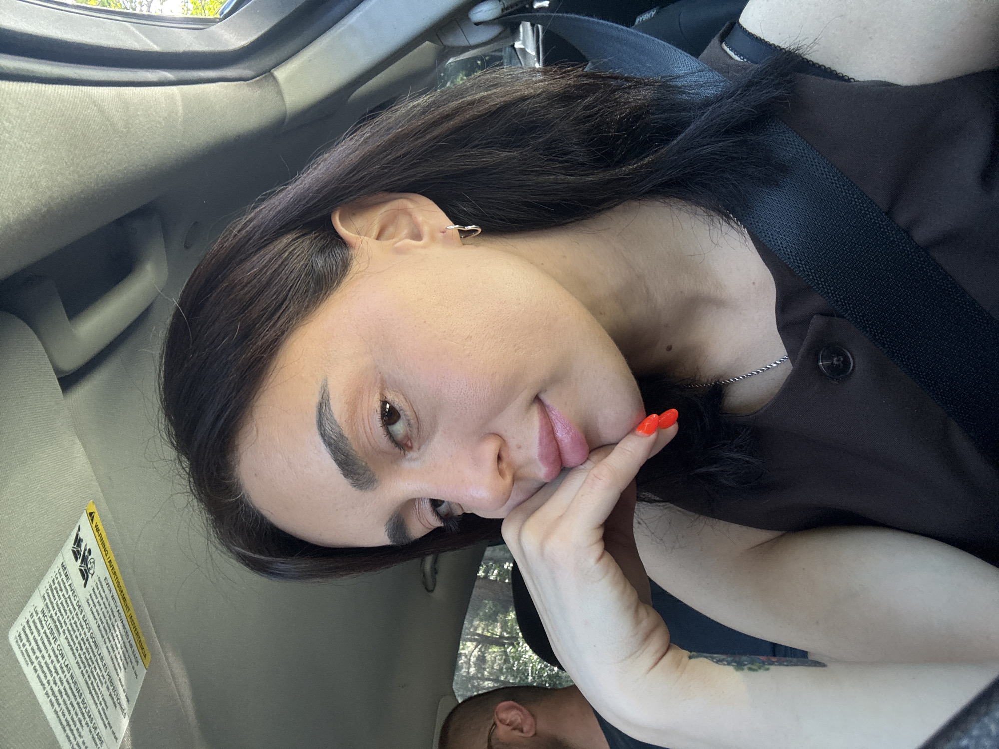 A woman is sitting in the passenger seat of a vehicle, with her left hand resting on her chin. She has dark hair, wears a brown top, and has bright orange nail polish. A seatbelt is visible across her chest. In the background, a man with a shaved head is visible. The vehicle appears to be driving through a wooded area, suggesting a location outside of a city center, such as on the outskirts of Tbilisi, Georgia.