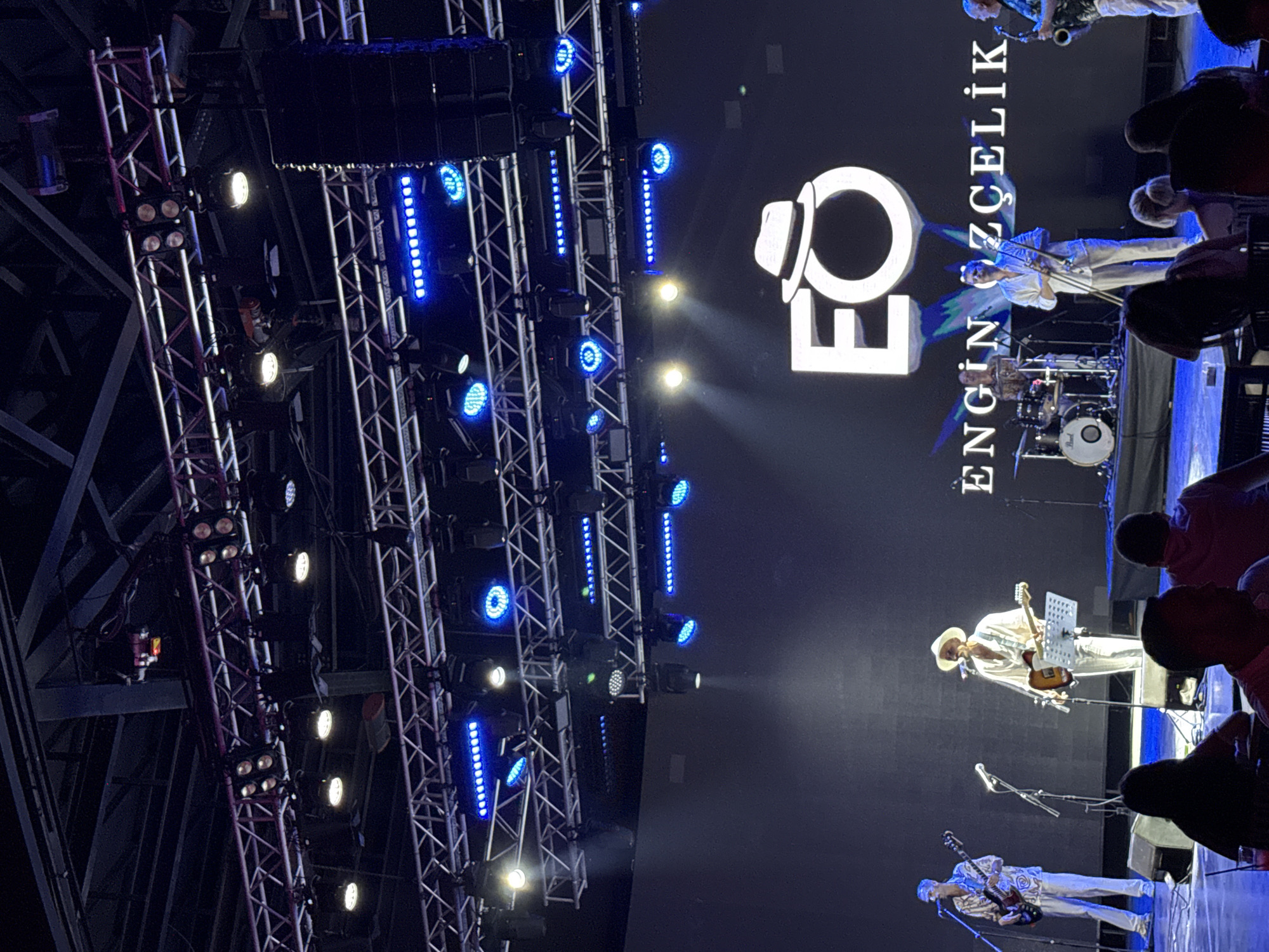 The image shows a live musical performance on a stage with a large screen displaying the logo "EO" and the text "ENGİN ÖZELİK". Two guitarists are visible on the left, one playing a bass guitar and the other an electric guitar. Another musician is playing a trombet in the center, and a saxophonist is on the right. A drummer is partially visible in the background. The stage is lit with blue and white lights. In the foreground, audience members are seated at tables, partially obscuring the view of the stage. The overall setting suggests an indoor concert venue in Bodrum, Turkey.