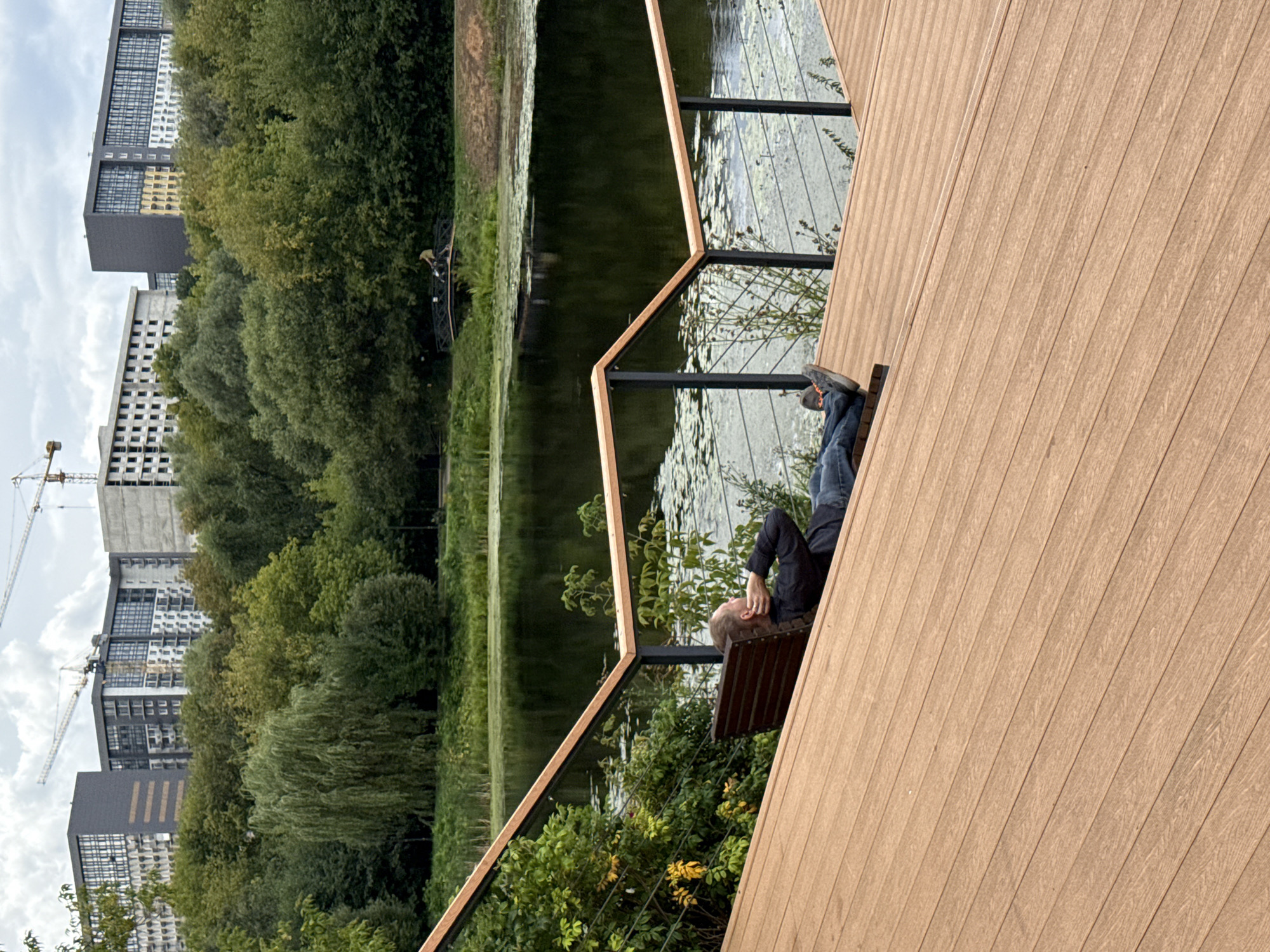 In Moscow, Russia, a man is seen reclining on a wooden deck overlooking a tranquil body of water. The deck, constructed with horizontal wooden planks, slopes downwards towards the water, featuring a metal railing with wooden accents. A dark wooden bench is placed on the deck, where the man is resting with his hands behind his head, his legs extended. He is wearing a dark long-sleeved shirt and blue jeans. The water, reflecting the overcast sky, is partially covered with lily pads. Beyond the water, a lush green forest with diverse trees forms a natural backdrop. In the distance, several multi-story residential buildings are under construction, with cranes visible against the sky, indicating ongoing development in the urban landscape. The scene captures a moment of relaxation amidst a developing cityscape and natural scenery.