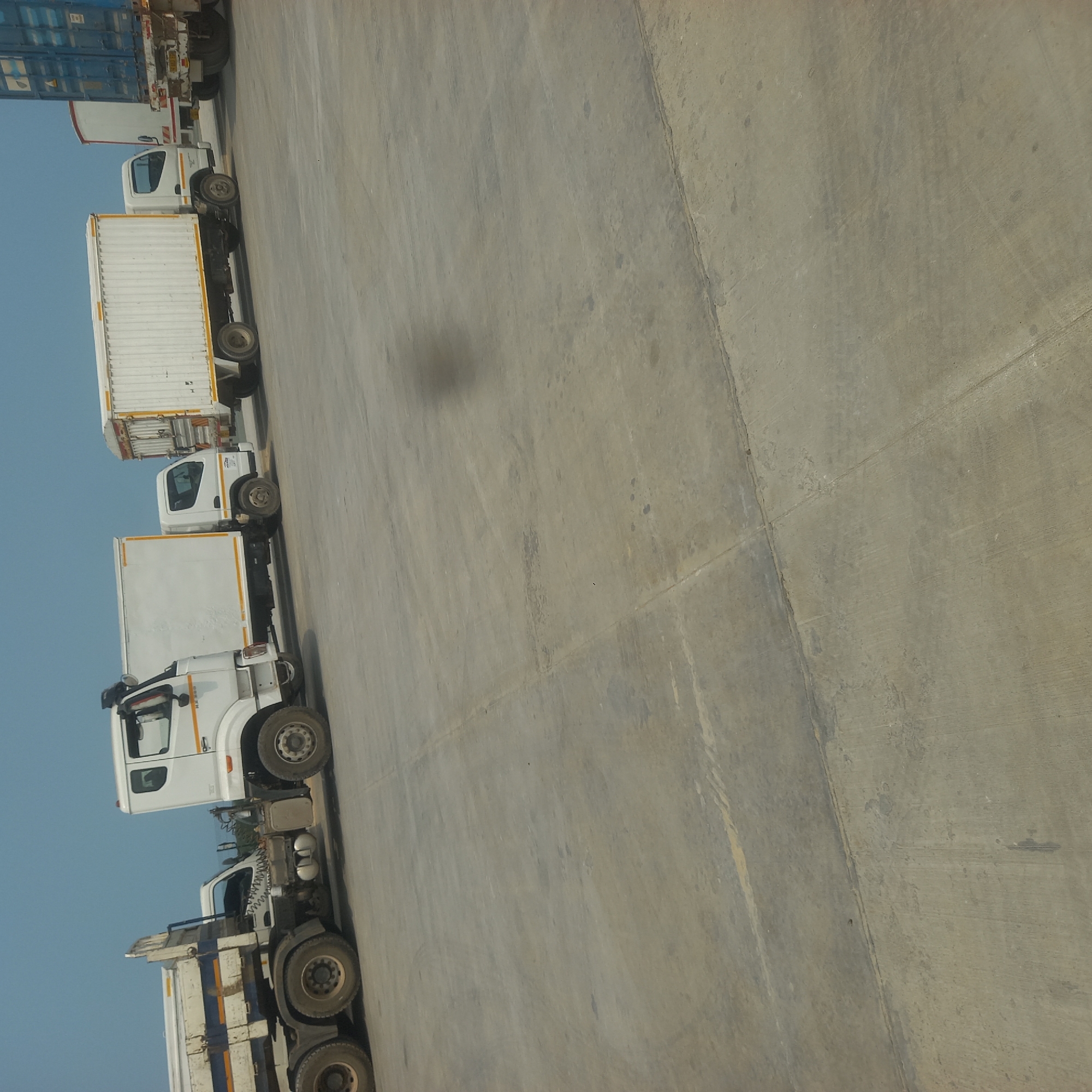 A line of trucks is parked on a concrete surface under a clear sky. The trucks are all white with some variations in their designs. The first truck on the left is a semi-trailer truck with its trailer appearing to be a tanker or a flatbed with a raised side. Next to it is a white box truck. Following that is another white box truck, and then a white refrigerated truck. The last visible truck is a semi-trailer with a large blue shipping container on its trailer. The ground is a worn concrete surface with visible tire marks and some darker stains. The scene appears to be a logistics or transportation hub. The location is Bububu, Tanzania.