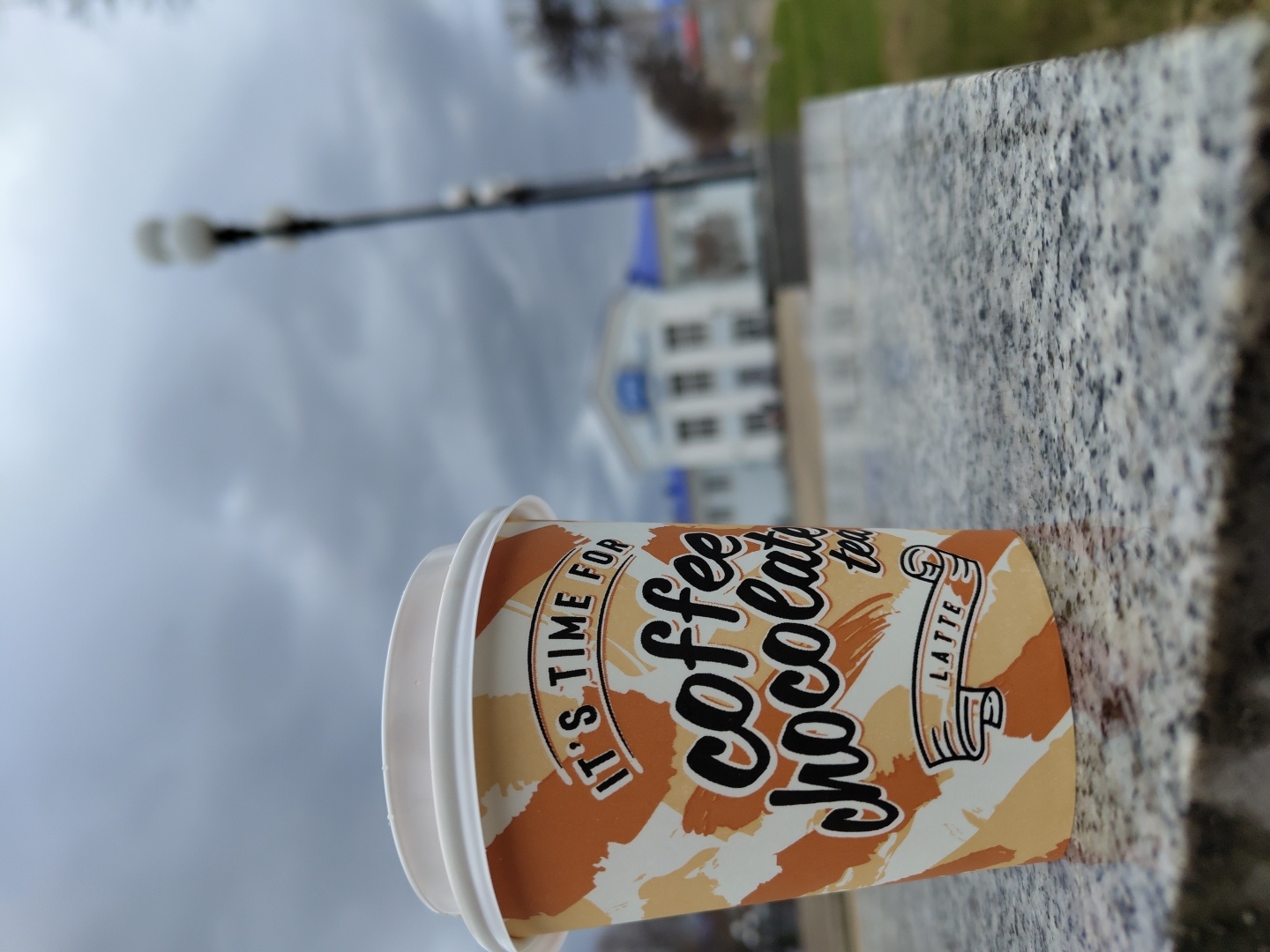 A disposable coffee cup rests on its side on a speckled grey and white granite surface, forming the central subject of the image. The cup features an alternating pattern of bold brown and light beige brushstrokes. Visible text on the cup reads, "IT'S TIME FOR," "coffee," "chocolate," "Latte," and "tea." The cup is sealed with a plain white plastic lid.
The background is significantly blurred, indicating a shallow depth of field. In the distance, an urban or town environment is visible under an overcast, light grey sky. A tall, dark street light pole with several luminaires extends upwards on the left side of the frame. Further back, a white building with blue roof details or trim is faintly discernible. The weather appears cloudy or overcast, consistent with daytime conditions. The scene suggests an outdoor setting, possibly a public area like a park or plaza in Chaikovsky, Russia, where the cup has been temporarily placed on a stone structure. No individuals are present in the image.