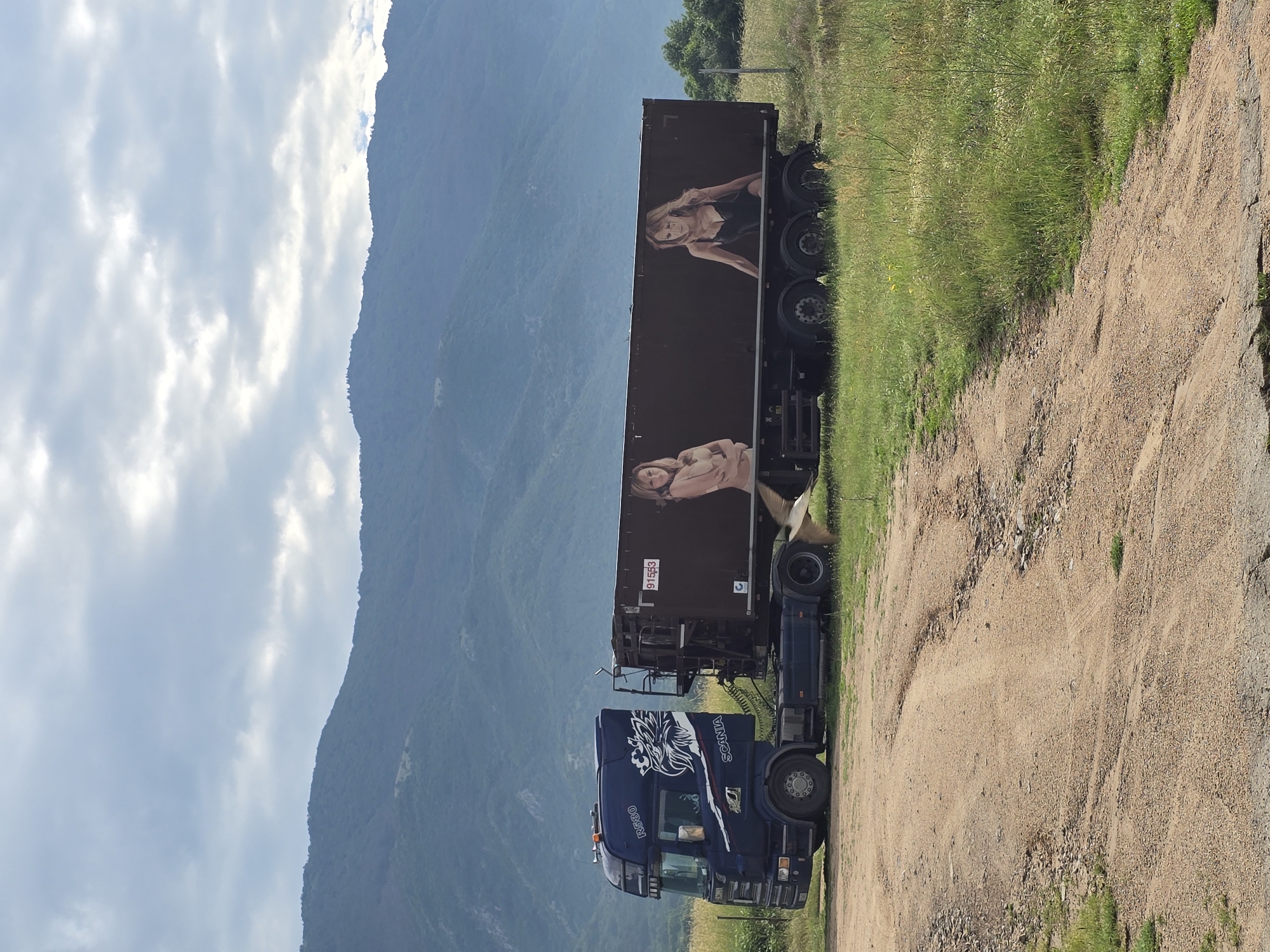 A blue Scania R580 truck is parked on a dirt path in a grassy field in Karnare, Bulgaria. The truck's cab features a white graphic of an eagle-like creature and the word "SCANIA" on its side. Connected to the truck is a dark brown trailer, which displays two large painted images of women. The woman on the left is depicted nude from the waist up, with her arms crossed, while the woman on the right wears a black low-cut top. A red rectangular label with the white numbers "91553" is visible on the upper left side of the trailer. The ground consists of dry, light brown dirt with tire tracks and patches of green grass and weeds. In the background, prominent dark green, tree-covered mountains rise under a partly cloudy, light blue sky. A small, light-colored bird is captured in flight near the lower right side of the trailer.