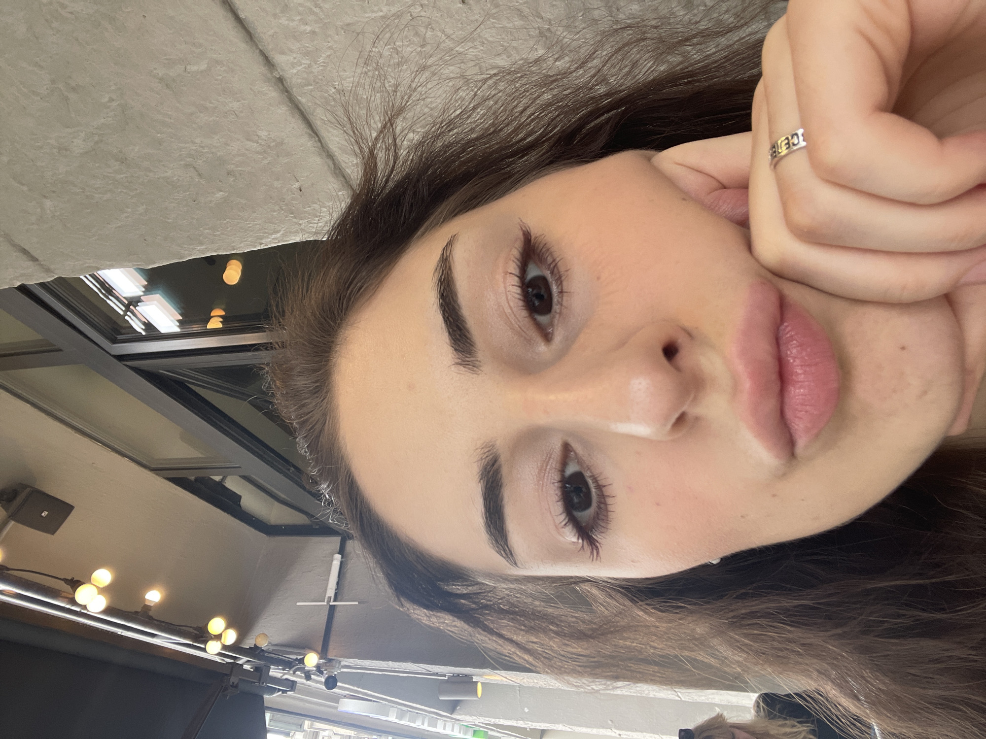 A young woman with long brown hair is seen in a selfie. She has fair skin, defined eyebrows, and dark eyes. Her lips are full and slightly parted. She is wearing a silver ring on her hand, which is propping up her face. In the blurred background, there's a ceiling with overhead lighting and a window structure. The location is suggested to be in Moscow, Russia.
