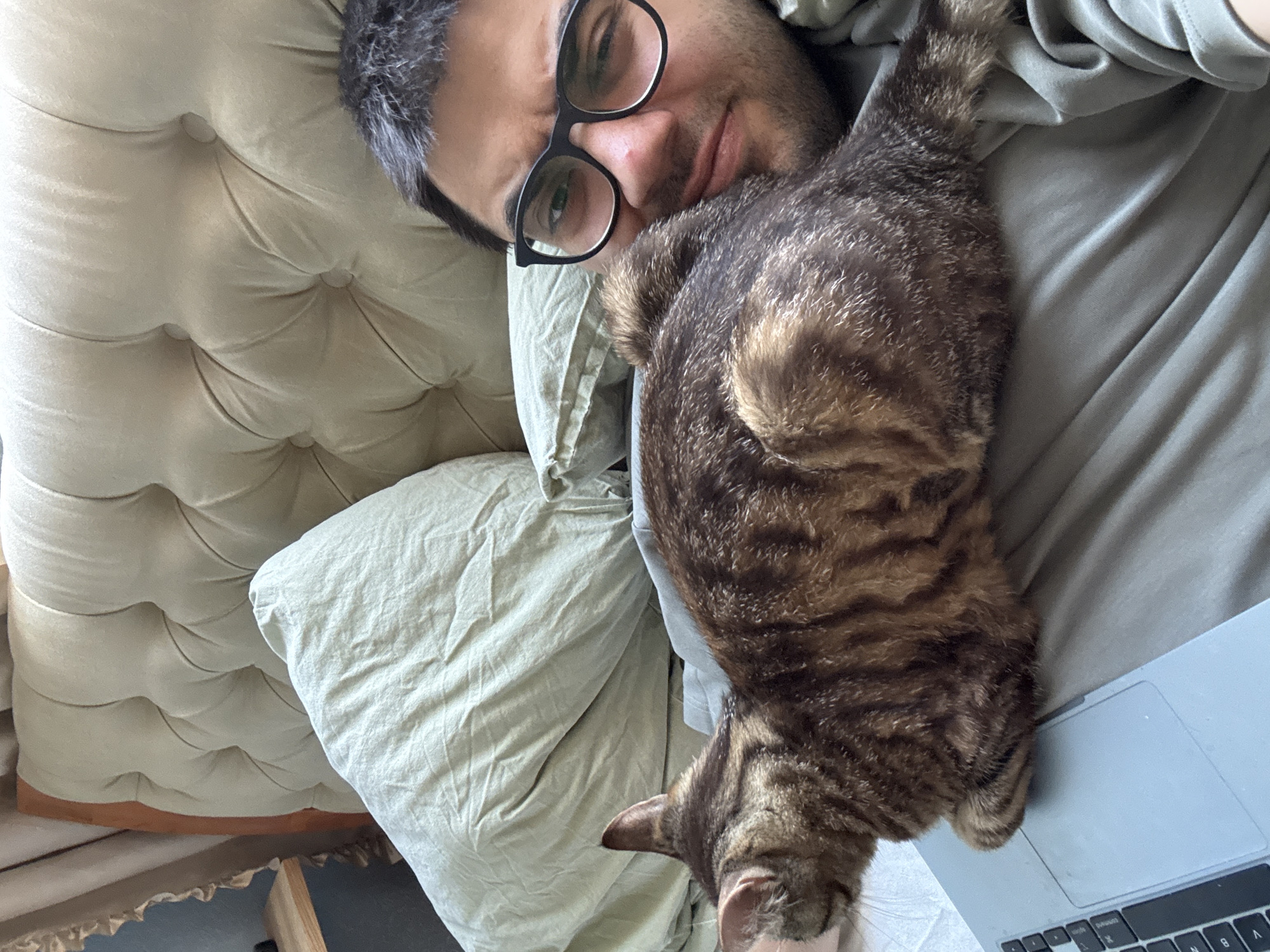 A close-up, overhead shot shows a person with glasses lying down with a tabby cat on their chest. The person is smiling and looking at the camera. The cat is curled up on the person's chest, with its head resting near the person's face. The background consists of bedding and a tufted headboard. A laptop is visible on the right side of the frame, partially covered by the cat. The lighting suggests it is daytime. The image conveys a sense of comfort and companionship. No visible text is present in the image. The context provided indicates the location as Bila Tserkva, Ukraine.