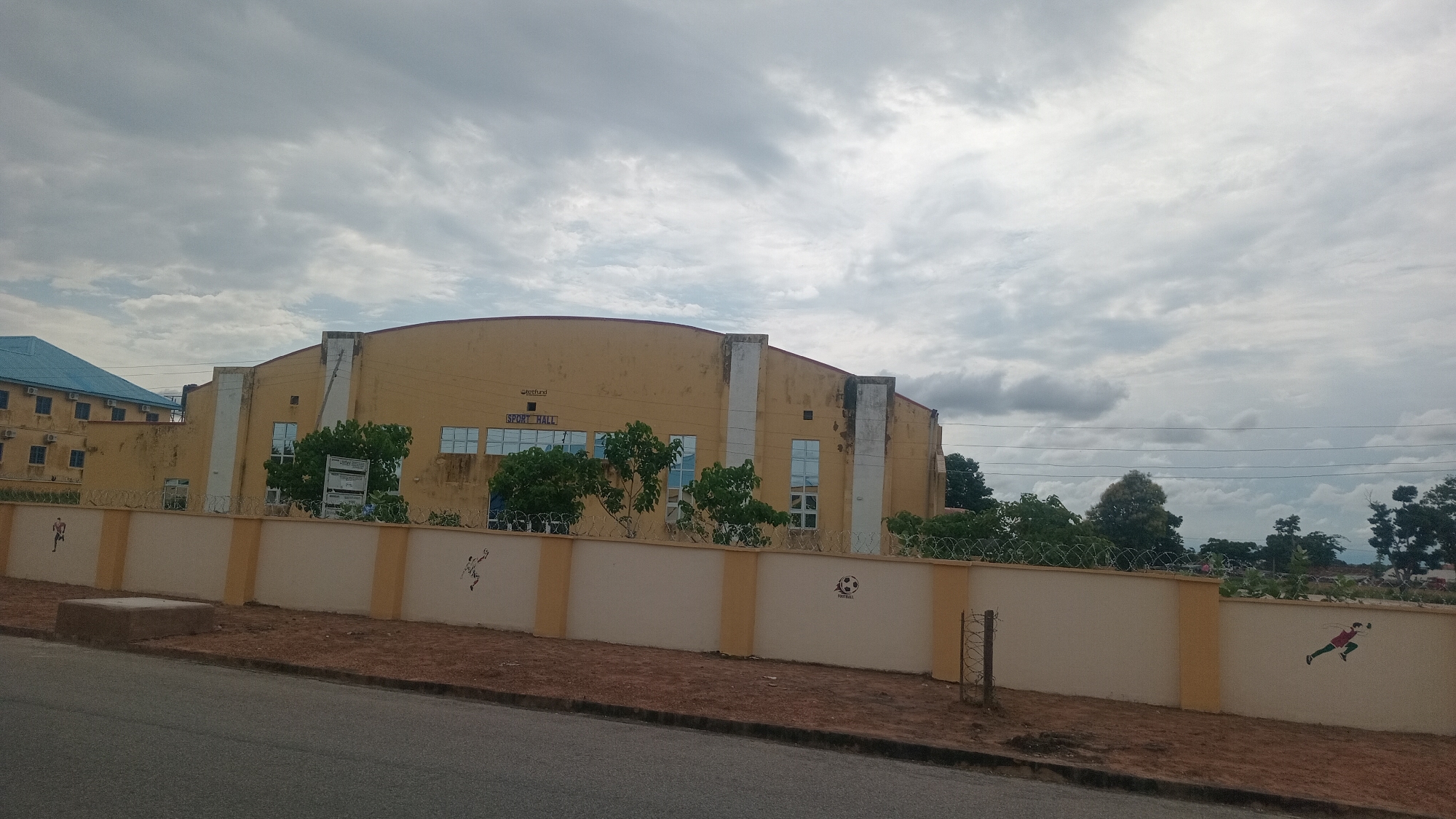 The image shows a tan-colored building with a curved roof labeled "SPORT HALL" located in Jalingo, Nigeria. The building has several large windows with blue frames. In front of the building is a beige-colored wall adorned with artwork depicting athletes. The wall is topped with barbed wire. Trees and other vegetation are visible behind the wall and around the building. The sky is cloudy. To the left of the "SPORT HALL" is another building with a blue roof. The scene is viewed from a street with a paved surface.