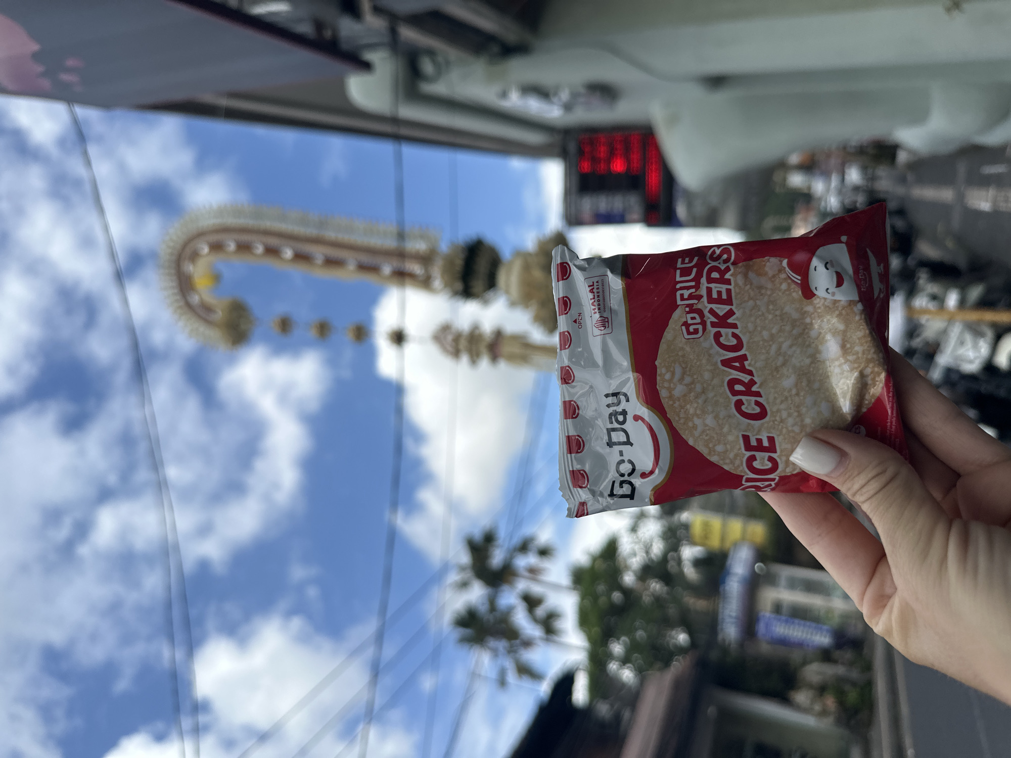 In the city of Ubud District, Indonesia, a hand holds a bag of Go-Day Rice Crackers. The bag is red and white, featuring the brand name "Go-Day" and the words "RICE CRACKERS". A cartoon character is visible on the lower right corner of the bag. In the background, a tall, decorative structure with lights is visible against a blue sky with white clouds. Palm trees and buildings line a street, where motorcycles are parked. The overall scene suggests an outdoor urban environment.