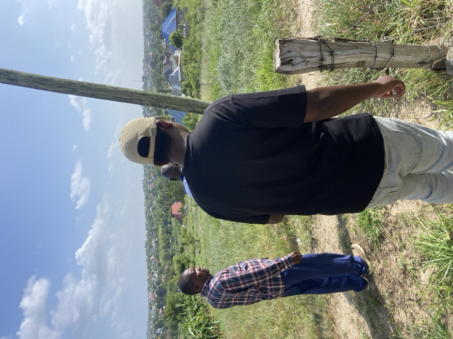 The image captures an outdoor scene in Kibaha, Tanzania, under bright daylight. Two men are prominently featured, standing on a dirt path overlooking a wide landscape.
In the foreground, a man is seen from a rear-side perspective, wearing a black t-shirt, a light tan baseball cap, and light blue jeans. His right arm hangs loosely by his side. Further into the middle ground and to the left of the foreground figure, a second man is visible, standing and facing towards the left (viewer's right). He is dressed in a plaid shirt with red, blue, and white patterns, dark blue trousers, and light brown shoes. A third individual's head is partially visible behind the foreground man, suggesting another person is present.
The men appear to be observing the expansive view before them, which consists of a valley or hilly area densely covered with green foliage and numerous houses. The houses display various roof colors, including blue, red, and grey, scattered amongst the trees.
The immediate surroundings feature a dirt path, green grass, and natural vegetation. On the left side of the image, a tall wooden utility pole stands vertically. To the right, a weathered wooden post, wrapped with barbed wire, is embedded in the ground.
The weather is sunny with a clear sky dotted with white clouds, indicative of daytime, likely morning or early afternoon. No visible text is present in the image. The overall scene suggests a quiet moment of observation in a developing rural or semi-urban setting.
