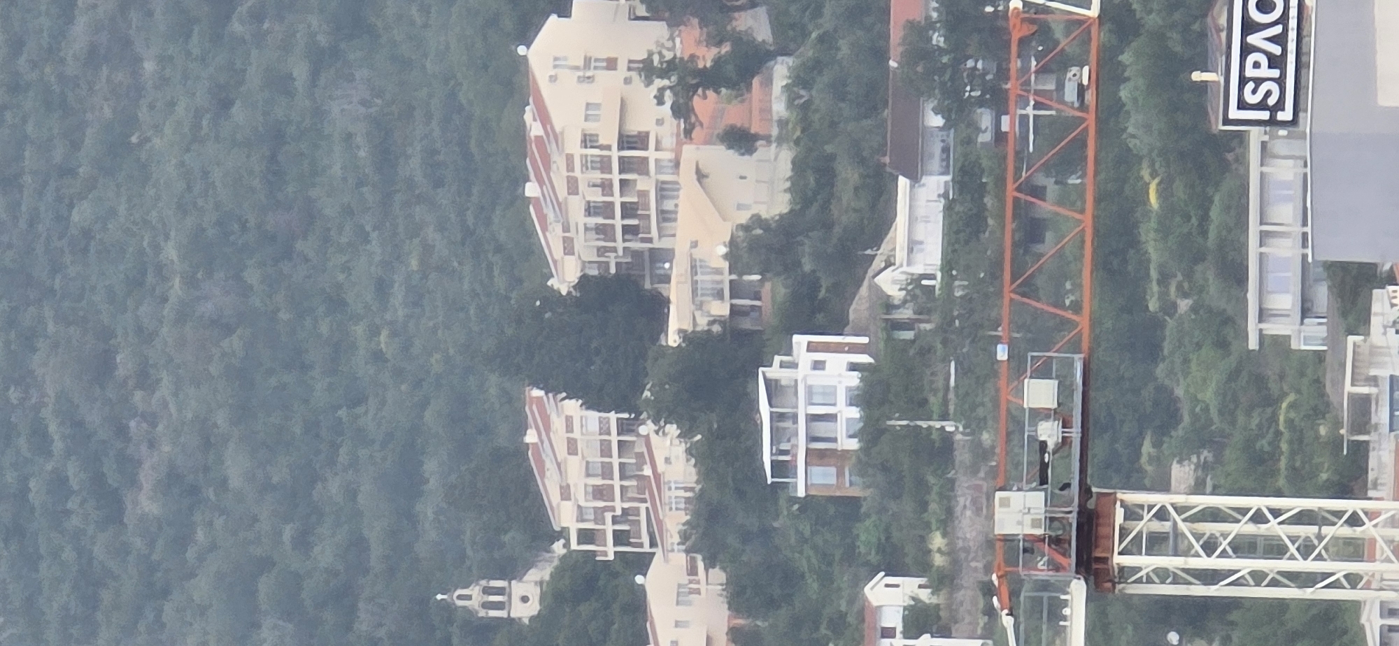 The image presents an aerial or elevated view of a coastal or hillside settlement, characterized by dense vegetation and various buildings, with ongoing construction. The scene appears to be under an overcast sky or during a hazy daytime, resulting in soft, diffused lighting and muted colors.
On the left side of the frame, a sprawling, densely forested hillside dominates, covered in lush green trees. On the right, a cluster of residential buildings and structures is visible, interspersed with trees. These buildings vary in architectural style; some are multi-story apartment complexes with light-colored facades and reddish-brown tiled roofs, featuring numerous balconies. Others appear more modern with white or light grey finishes, large windows, and simpler designs.
A prominent orange and white construction crane stands tall in the mid-right portion of the image, signifying active development in the area. Near the crane, on a building, a rectangular black sign is affixed vertically, clearly displaying the word "SPACE" in white, sans-serif capital letters. The overall setting suggests a growing community nestled within a natural, green environment, likely in a coastal or hilly region like Bar, Montenegro. No people are visible.