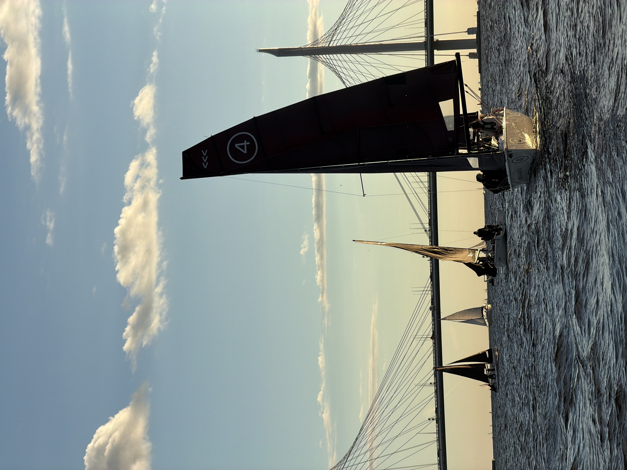 The image shows a fleet of sailboats on the water, with a cable-stayed bridge in the background. The sky is blue with scattered clouds, and the sun appears to be setting, casting a warm glow on the scene. The water is choppy, indicating a moderate breeze.
In the foreground, a white sailboat with a dark sail is prominently featured. It has a number "4" in a white circle on its sail. Several people are visible on board, appearing to be racing or sailing.
Further in the background, other sailboats are visible, some with dark sails and others with lighter sails. The cable-stayed bridge spans the water, with its towers and cables creating a distinctive silhouette against the sky. This bridge is likely part of the Western High-Speed Diameter, a major transportation artery in Saint Petersburg, Russia. The presence of the bridge and the Neva River or Gulf of Finland waters suggest the location is within or near Saint Petersburg. The overall atmosphere is one of a sporting event or recreational activity taking place on the water during the golden hour.
