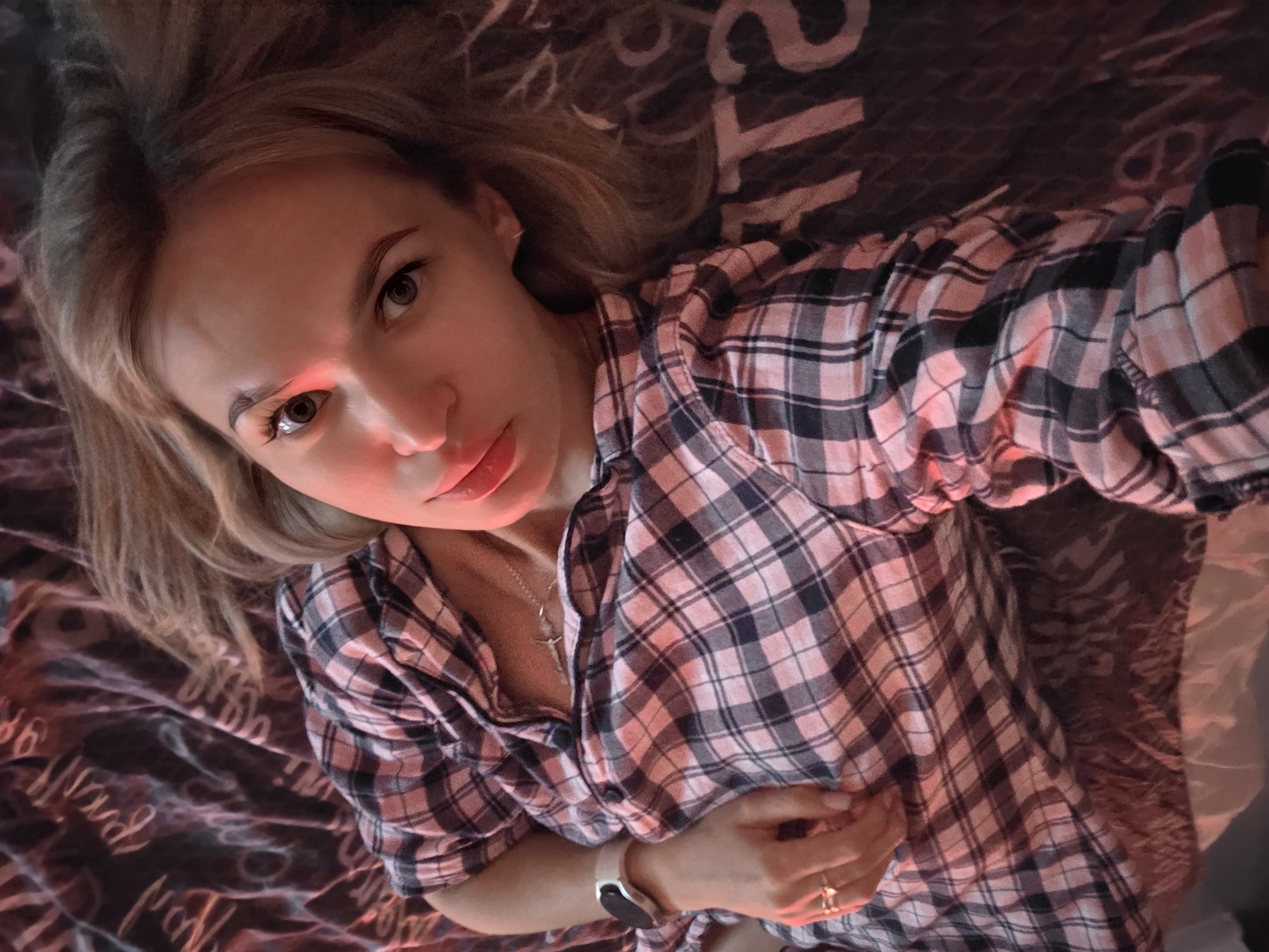 A woman lies on a bed in what appears to be an indoor setting. She has fair skin, light blonde hair, and is wearing a plaid button-down shirt. She is wearing a smart watch on her left wrist and a ring on her right hand. A necklace is visible around her neck. The bed has a patterned bedspread, and the lighting casts a red hue over the scene. The location is likely inside a residential building in the city of Chaikovsky, Russia.