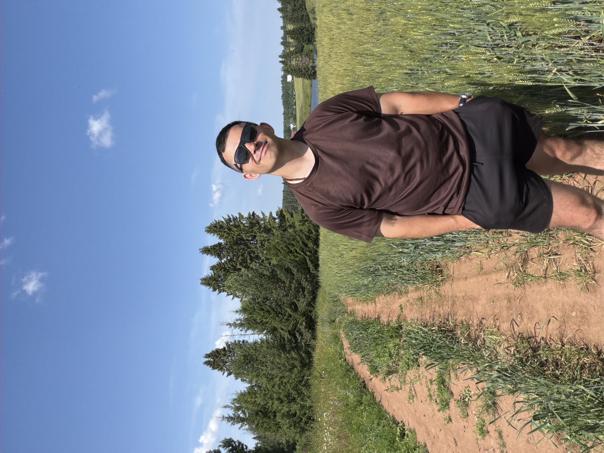 A medium shot captures a man standing on a dirt path in a rural landscape. He is wearing sunglasses, a brown t-shirt, and black shorts. The path runs through a field of tall green grass, with a dense forest of evergreen trees visible in the background. The sky is a clear blue with a few scattered white clouds. The location is identified as Podshivalovo, Russia.
