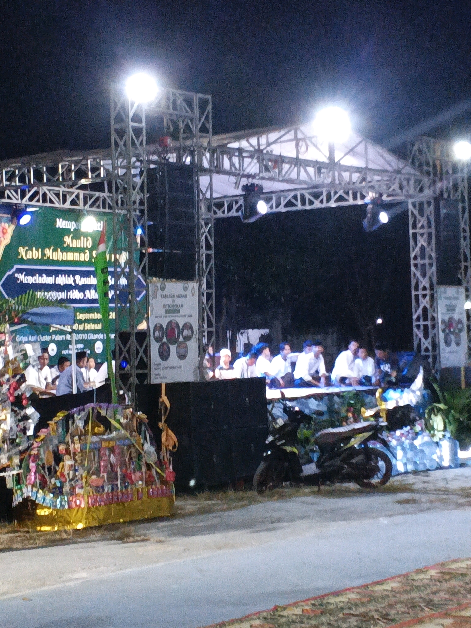 The image depicts an outdoor nighttime event in Panamping, Indonesia, likely a religious ceremony or festival. A large stage structure made of scaffolding is prominently featured, illuminated by bright lights. In front of the stage, a group of people are seated, appearing to be an audience or participants. To the left of the stage, a green banner with text is visible, along with a colorful, decorated float or display. To the right of the stage, a sign with circular images is posted. In the foreground, a motorcycle is parked on the side of a road. The overall scene suggests a community gathering or celebration taking place after dark.