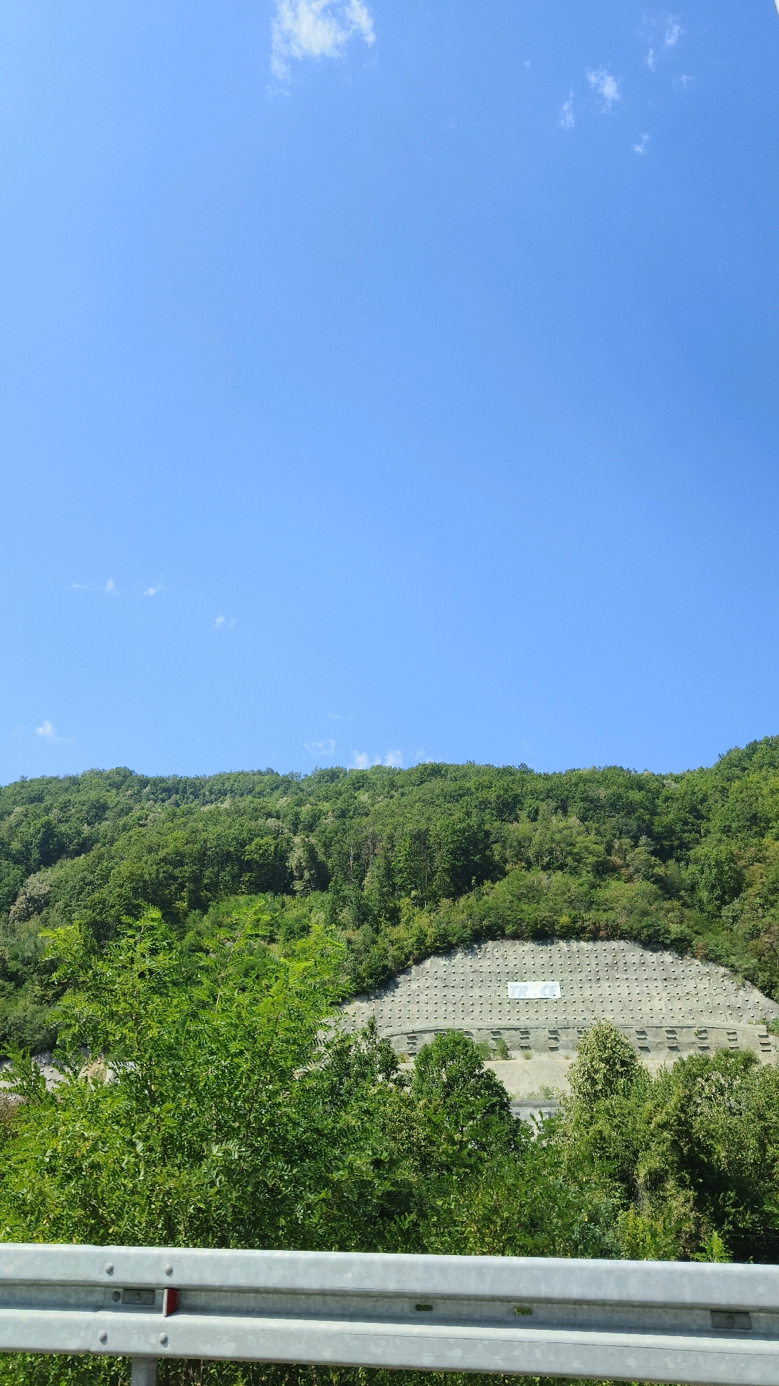 The image shows a green, tree-covered hillside under a clear blue sky with a few wispy clouds. In the foreground, a metallic guardrail is visible, suggesting a road or viewing area. On the hillside, a man-made structure, possibly a retaining wall or stabilization project, is evident, with rows of concrete anchors and a white banner displaying the text "TR.CE". The setting appears to be a natural, somewhat rugged landscape.