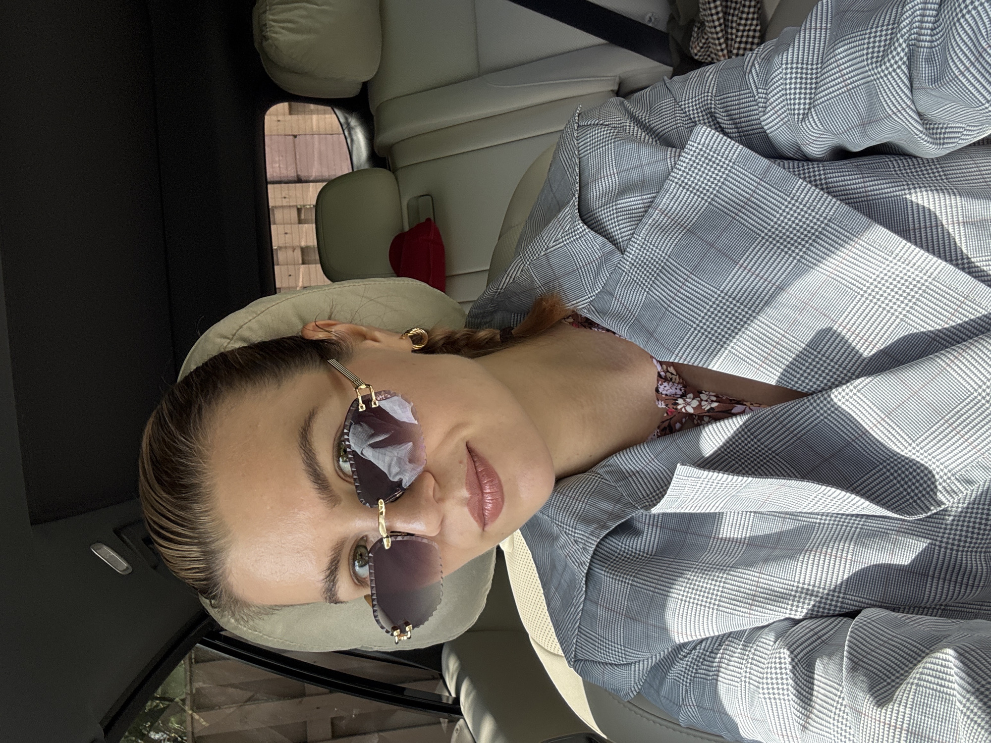 A woman with slicked-back hair is seated in a car wearing sunglasses and a gray plaid blazer. She is wearing a seatbelt. The interior of the car is light gray leather. The location is unknown.