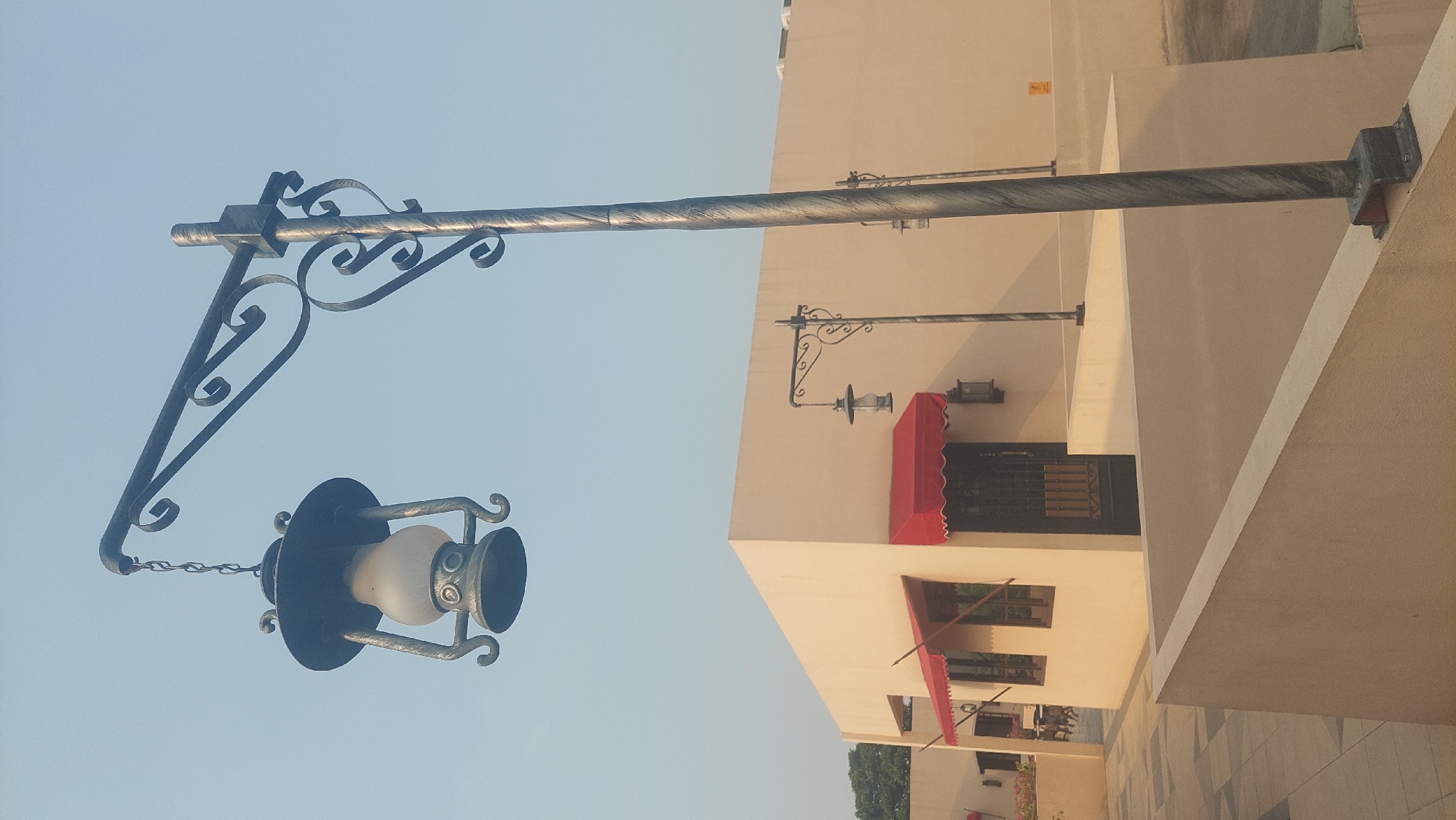 The image depicts an outdoor scene in Cileungsi, Indonesia. A large, ornate streetlamp with a hanging lantern stands prominently in the foreground, its metalwork featuring decorative scrolls. Behind it, a light-colored building with red awnings over windows and a black, barred door is visible. Several smaller, similar streetlamps are placed at intervals along the building's exterior. The environment appears to be a courtyard or walkway, with a light-colored tiled floor in the lower left and a low, beige wall in the foreground. The sky is a clear, pale blue, suggesting it is daytime. There are no people visible in the image.