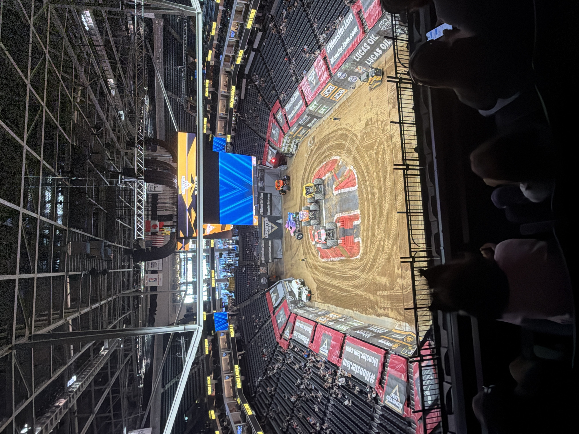 This image, taken from a high vantage point within an arena, depicts a monster truck event. The arena, likely the Golden 1 Center in Sacramento, California, features tiered seating filled with spectators. In the center of the arena floor, a dirt track has been set up for the event, with several monster trucks, including a red one and an orange one, positioned on or near the track. A large screen displaying blue geometric patterns is visible at one end of the arena. The overall scene suggests an indoor sporting event or entertainment show.