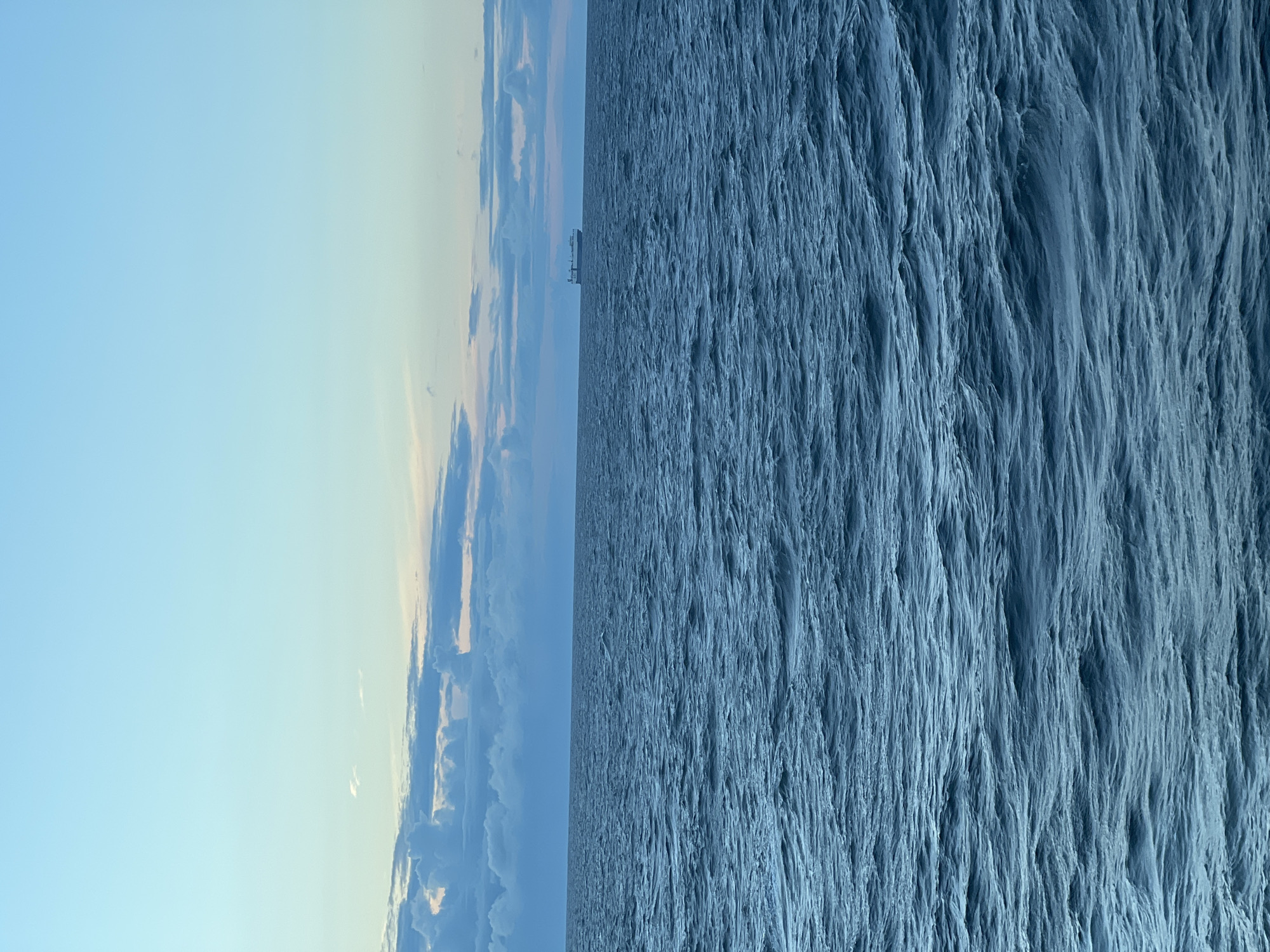 A wide shot captures a serene ocean under a pale blue sky. The water is a deep, dark blue with subtle ripples, indicating a calm sea. In the distance, a large white ship is visible on the horizon, a solitary object against the vast expanse of water. The sky transitions from a light blue at the top to a softer pinkish hue near the horizon, where scattered clouds add texture. The overall impression is one of tranquility and open space. The location is Unknown, Poland.