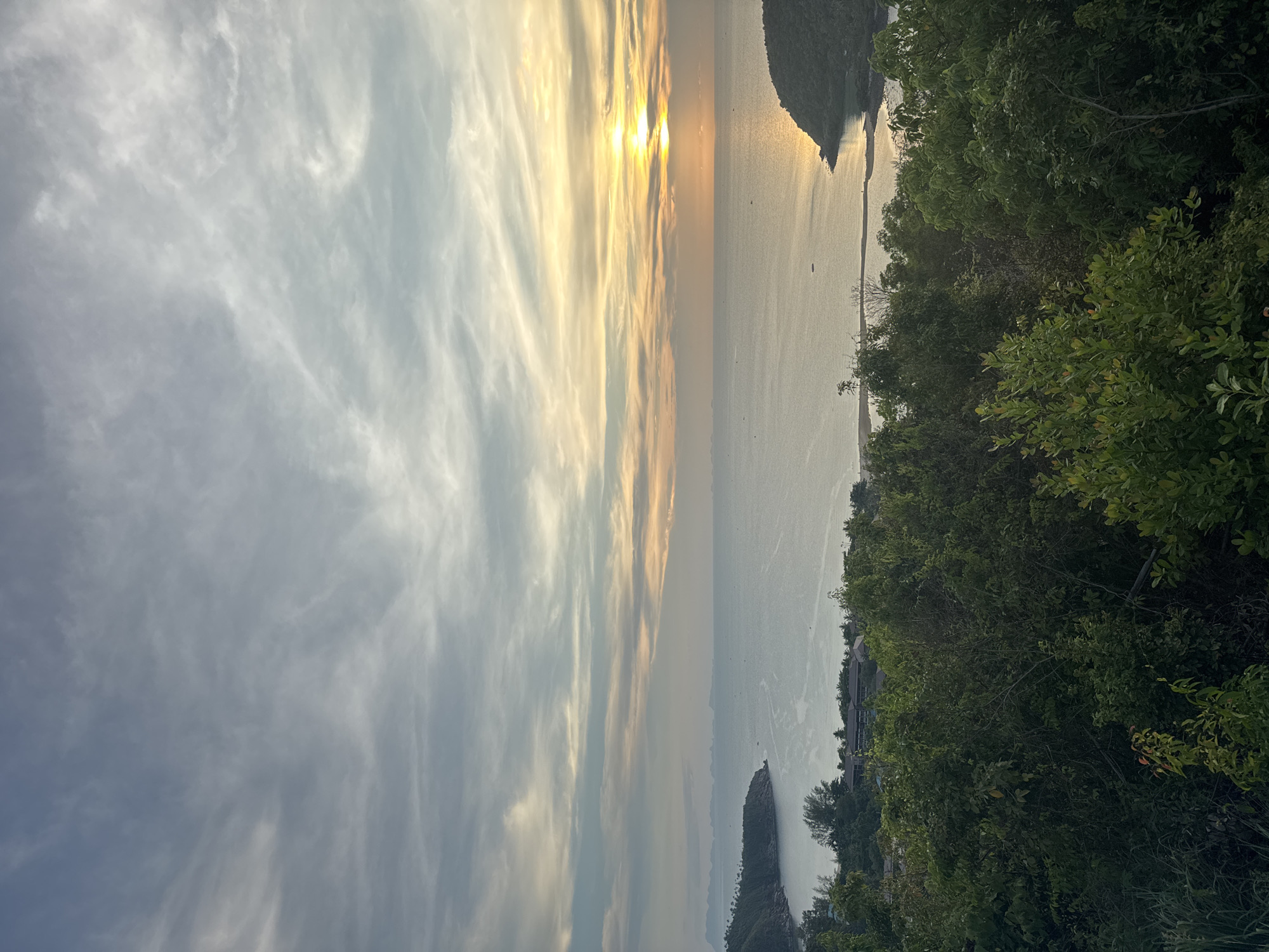 The image shows a vertical view of Baan Wang Ta Kien, Thailand, at sunset. The sky, filled with wispy clouds, transitions from a bright, sunlit area near the horizon to a darker shade of gray. The sun's golden light reflects on the calm water surface. Green foliage occupies the right side of the image, bordering the sea. In the distance, land masses appear along the horizon.