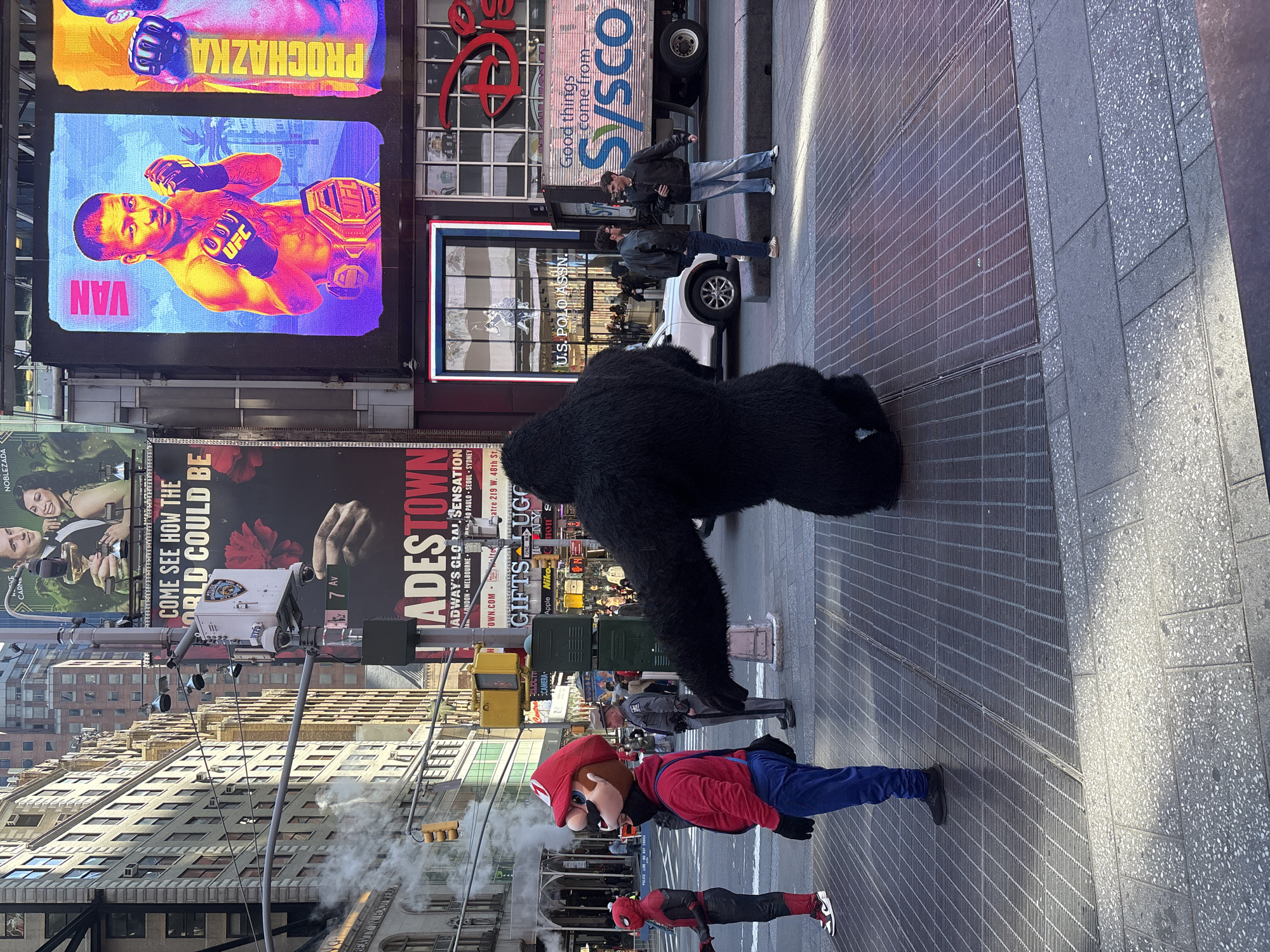 The image depicts a street scene in Times Square, New York City. A person in a Mario costume stands on the sidewalk, next to a person dressed as Spiderman. Another individual in a gorilla suit is positioned further up the street. The background is filled with bright billboards and signs, including advertisements for UFC and the musical "Hadestown". Buildings are visible behind the signs. The street appears to be a pedestrian zone, with people walking in the background and cars in the distance. Steam rises from a grate in the street.