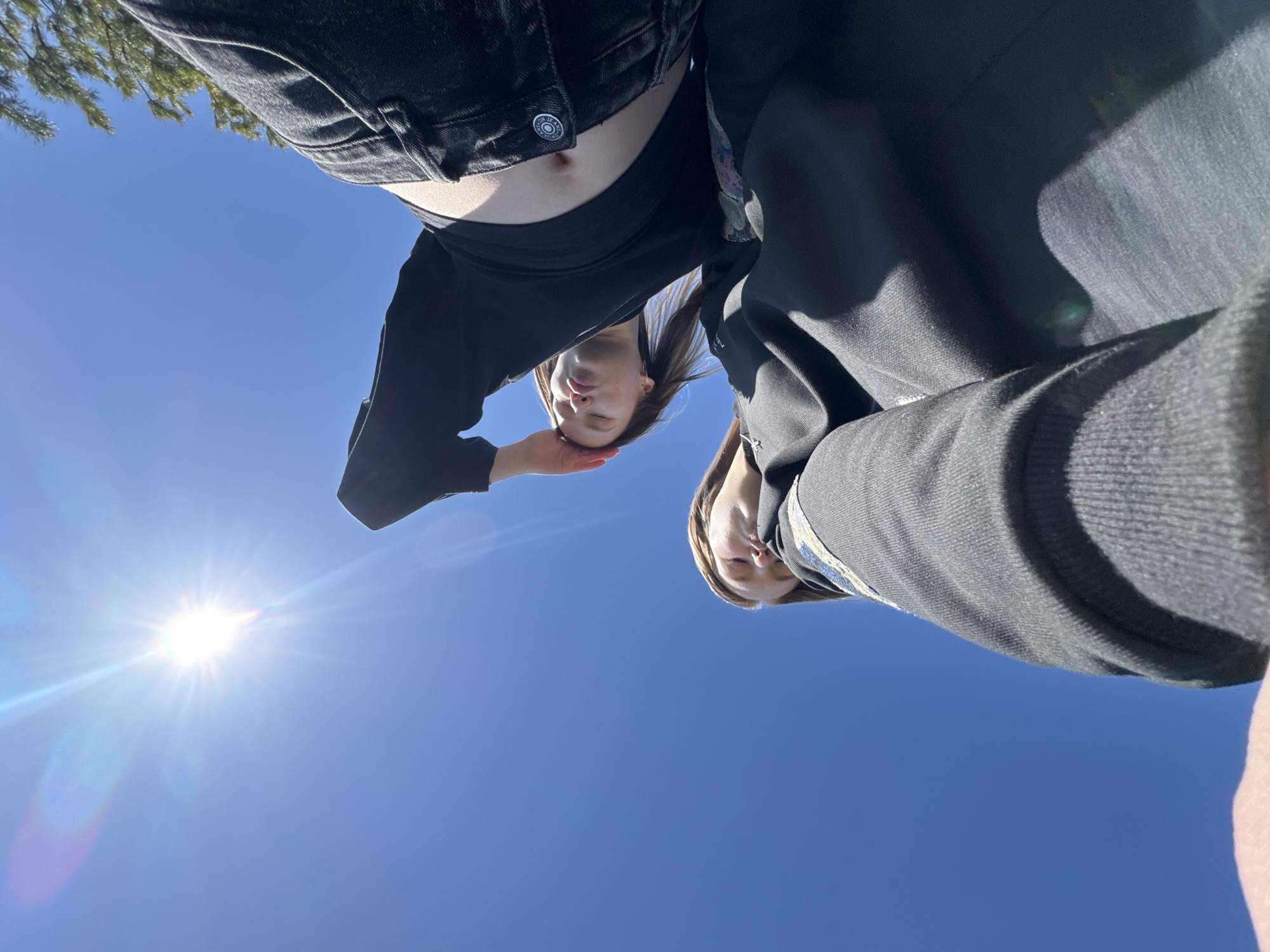 The image is a low-angle, upward-facing shot of two individuals against a bright blue sky with the sun visible in the lower left corner. The subject on the left is wearing black jeans and a black long-sleeve shirt, exposing a small portion of their midriff. Their face is visible, and they appear to be looking down. To their right is another person, wearing a dark hooded sweatshirt. Only a portion of their face is visible. The background consists solely of a clear blue sky and the bright sun. There is no indication of the location being in Zhigalki, Russia.