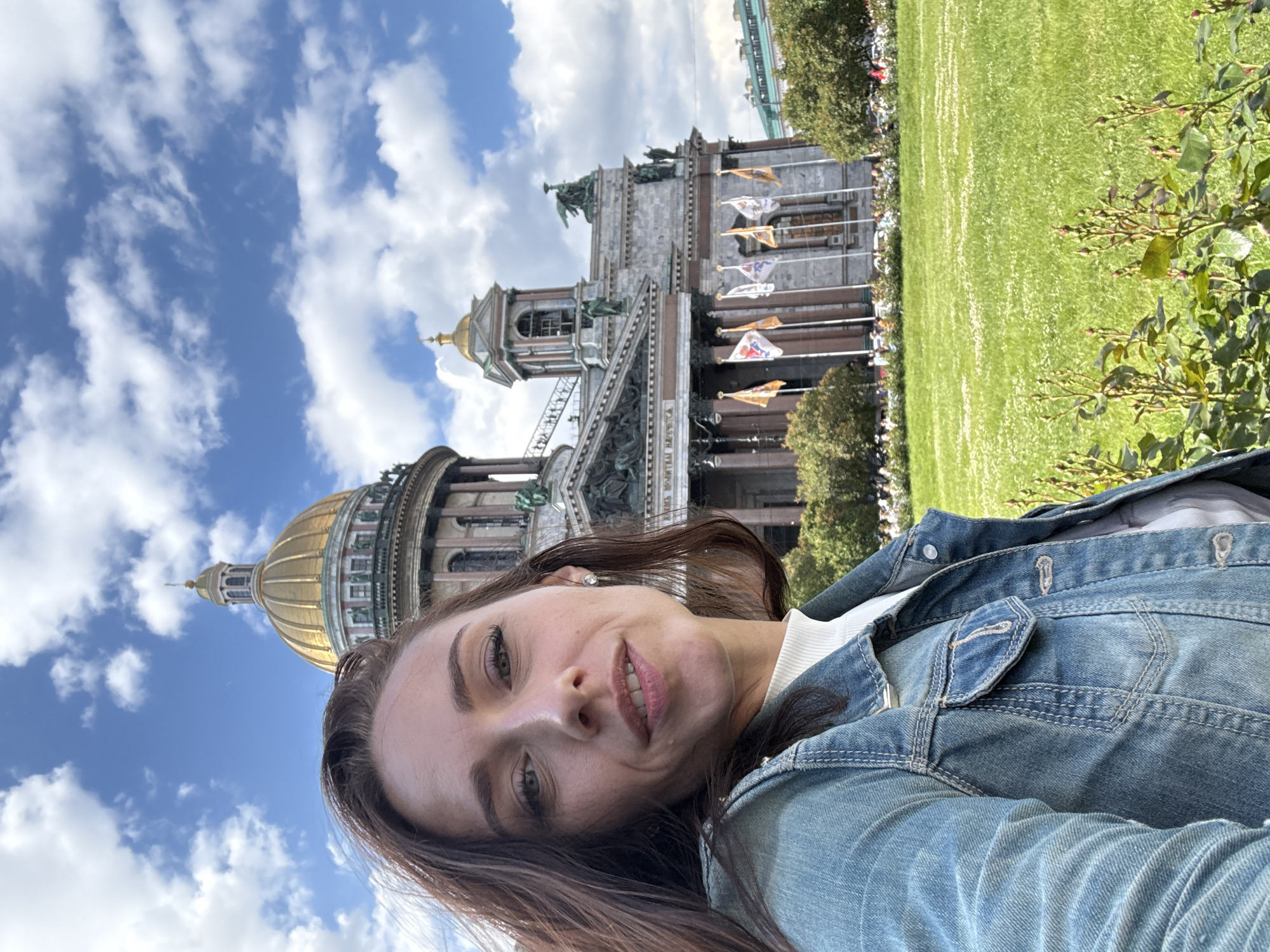 A woman with long brown hair, wearing a denim jacket over a white turtleneck, is taking a selfie in front of St. Isaac's Cathedral in Saint Petersburg, Russia. The cathedral, a prominent landmark, features a large golden dome and intricate architectural details. The woman is looking at the camera, smiling slightly, with the sunlit facade of the cathedral behind her. The scene is set on a bright, cloudy day, with a lush green lawn and trees in the foreground and middle ground, respectively. Several flags are visible on poles in front of the cathedral.