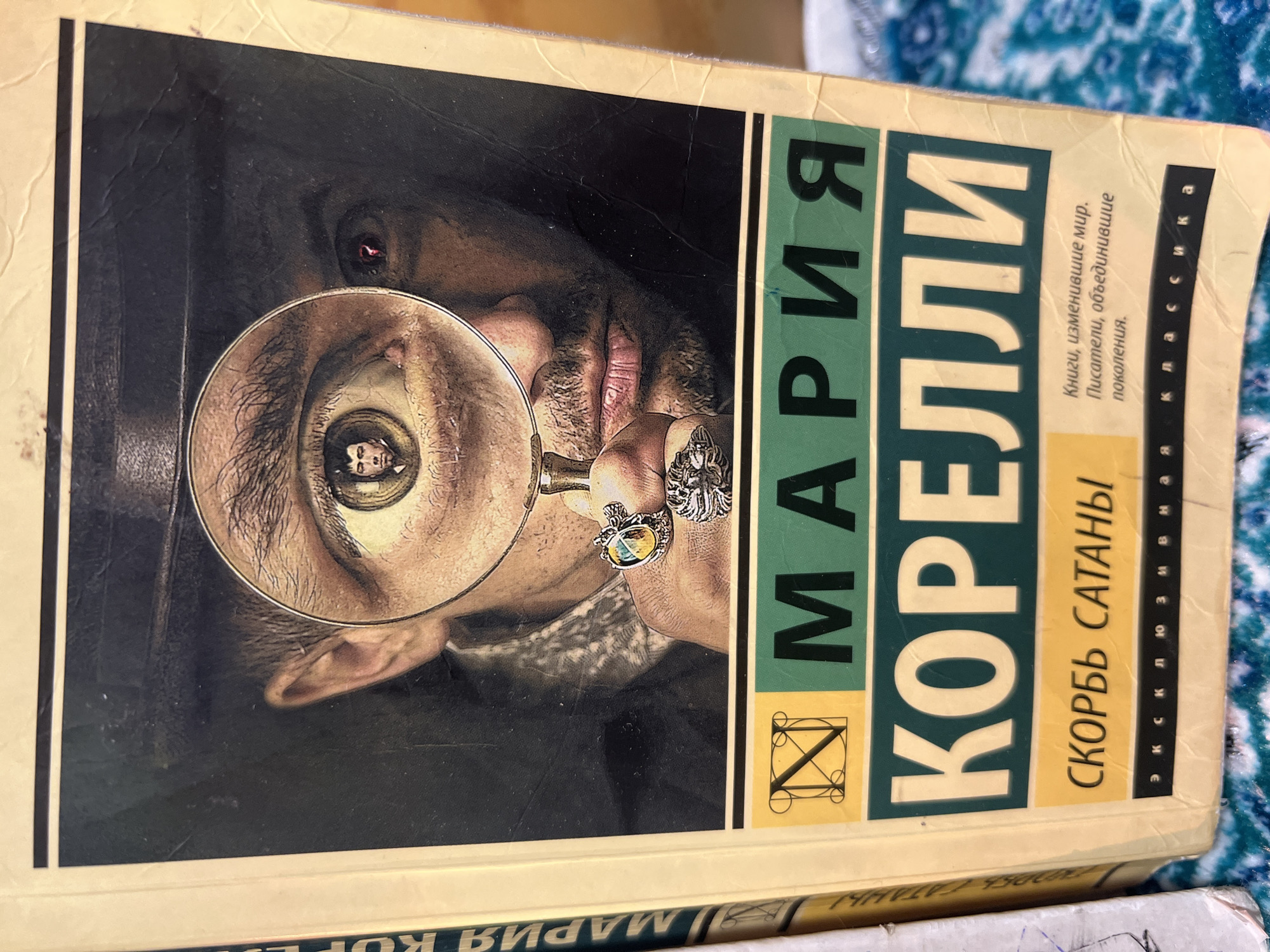 The image displays the cover of a book titled "The Sorrows of Satan" by Marie Corelli, in Russian. The book cover features a detailed illustration of a man with dark hair and a beard, wearing a hat, holding a magnifying glass to his right eye. The magnifying glass significantly enlarges his eye, and within the reflection of the lens, a smaller, contemplative face can be seen. The man wears two ornate rings on his right hand, which holds the magnifying glass. His left eye is partially visible in the upper right corner of the illustration.
Visible Russian text on the cover includes:
* Author: "МАРИЯ КОРЕЛЛИ" (Mariya Korelli / Marie Corelli)
* Title: "СКОРБЬ САТАНЫ" (Skorb' Satany / The Sorrows of Satan)
* Series: "Эксклюзивная классика" (Exclusive Classics)
* Tagline: "Книги, изменившие мир. Писатели, объединившие поколения." (Books that changed the world. Writers who united generations.)
The book cover is yellowish-beige and shows signs of wear, including creases and minor scuffs, particularly along the top edge. The book is resting on a patterned blue and white fabric, possibly a blanket or tablecloth. Part of another book's spine is visible underneath the depicted book. The scene appears to be indoors in Ильинская Поляна Снт, Russia.