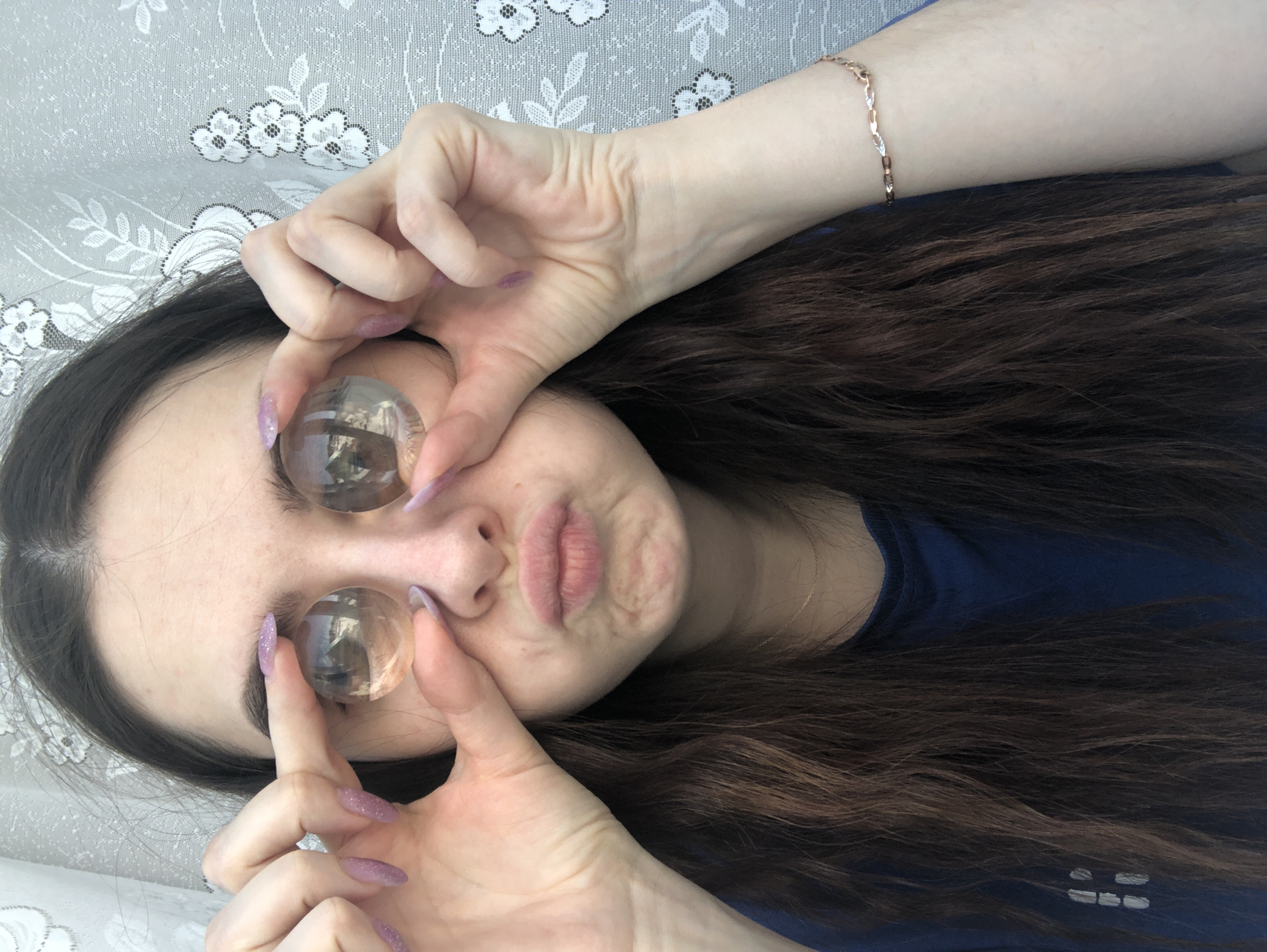 A person with dark hair and fair skin is lying on a surface with a floral pattern. They are wearing a blue top and a gold bracelet. The person is holding two transparent spheres over their eyes with their fingers. Their fingernails are long and painted a light purple color with glitter. The person's mouth is pursed.
The reflection within the spheres contains scenes of a city, specifically Noviy, Russia. The images are small and distorted, but buildings and urban infrastructure can be discerned.