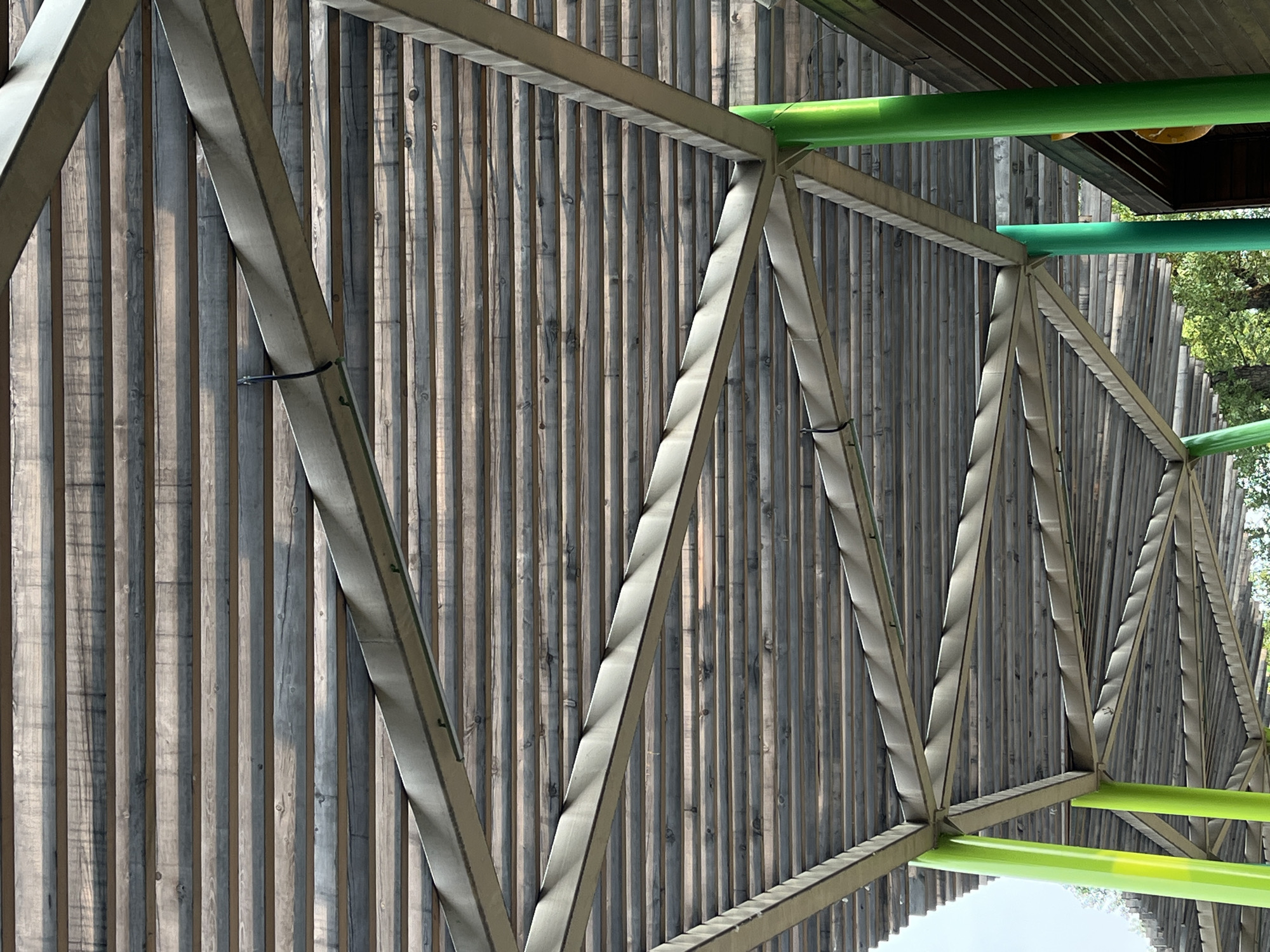 The image shows a view looking up at the underside of a wooden slatted roof supported by a metal framework and green pillars. The roof is constructed from many narrow, parallel wooden planks with visible grain and variations in color, suggesting they may be weathered. The metal framework is triangular and geometric, forming a repeating pattern above. Bright green cylindrical pillars support the structure, with some visible in the foreground and mid-ground. In the background, a brown wooden building is partially visible to the right, and the tops of trees can be seen against a light sky. The scene appears to be outdoors, possibly in a park or public area, and there are no people or animals visible in the image. The overall impression is one of modern architecture integrated with natural materials. The location in Krasnoyarsk, Russia is not discernible from the visual information provided in the image.