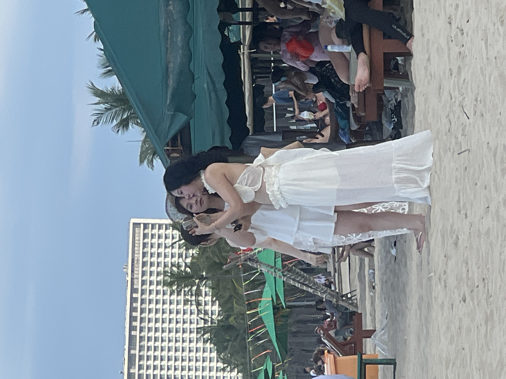 The image depicts a beach scene in Da Nang, Vietnam, with a large hotel visible in the background. In the foreground, three young women are standing on the sand. Two of the women are taking a selfie with a smartphone. They are all wearing white outfits. Behind them, beach umbrellas, lounge chairs, and other beachgoers can be seen. The overall atmosphere appears to be leisurely and social.