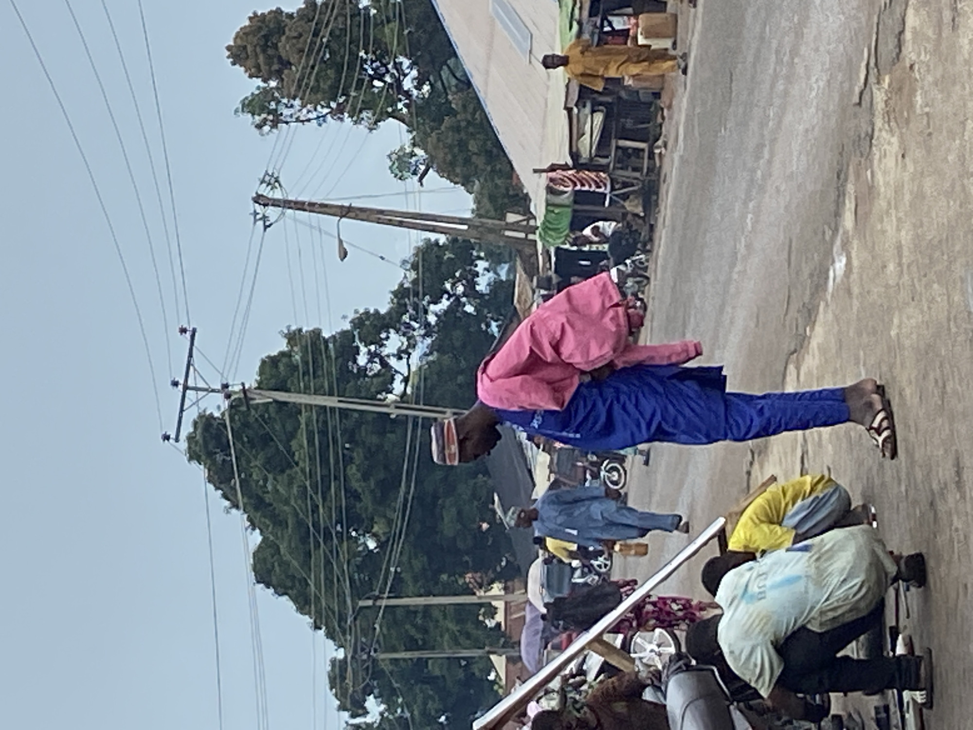 This image captures a daytime scene on an active street in Mararaba, Nigeria. The foreground shows an asphalt road, partially cracked, with a man in a light-colored t-shirt and dark trousers squatting on the left, seemingly engaged in work near the front wheel of a gray motorcycle or moped. To his right, a second individual in a yellow t-shirt and light blue pants is also squatting, facing away from the viewer. Slightly to the right and standing upright, a man wears a long blue tunic and pants with a bright pink garment draped over his left shoulder, exposing his dark sandals.
In the midground, various individuals are present. A man in a blue traditional garment walks toward the left, carrying a light-colored jug. Further back, an individual in a patterned dress is partially visible near a motorcycle. On the right, a man in a full yellow or light brown traditional outfit walks towards the right. Numerous informal market stalls line the right side of the road, constructed with corrugated metal roofs and wooden frames, displaying various goods. One stall shows colorful striped items, possibly fabrics. A roof on the far right midground features small solar panels. Several motorcycles are parked or being ridden by individuals throughout the midground.
The background is dominated by dense, green foliage from large trees. Multiple wooden and concrete utility poles, equipped with cross-arms and numerous power lines, crisscross the upper portion of the frame against an overcast, light blue sky. A street lamp is attached to one of the utility poles on the right. The overall impression is that of a bustling, semi-urban commercial area.