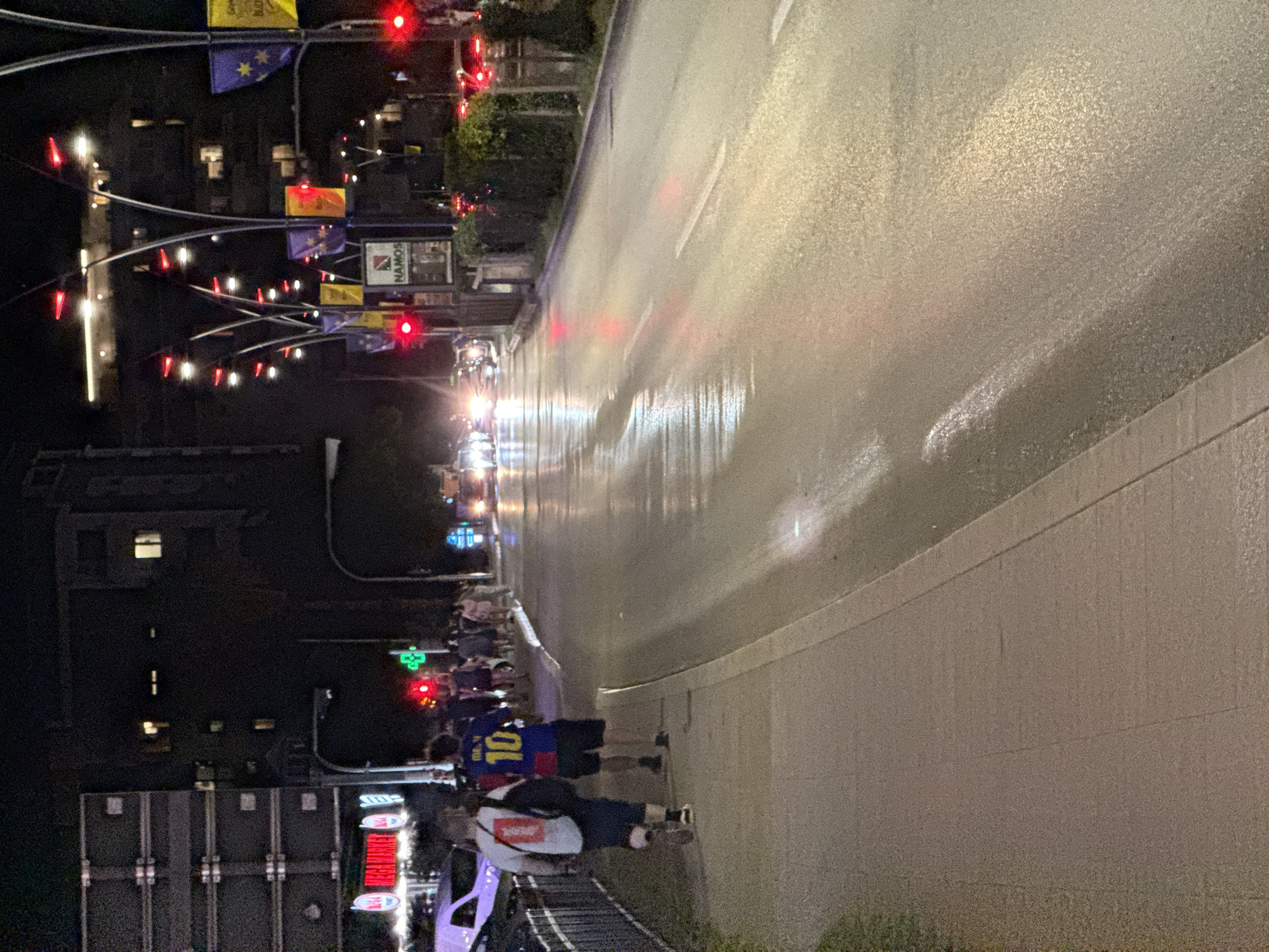 The image depicts a street scene at night in Budva, Montenegro. A paved sidewalk runs along the left side of the frame, where two individuals are walking away from the camera. The person in front is wearing a white t-shirt and dark shorts, carrying a backpack. The person behind them is wearing a blue and yellow jersey with the number "10" and dark shorts. Several other pedestrians are visible further down the sidewalk, moving towards the background.
The street itself is wet and reflects the lights from street lamps, traffic signals, and vehicles. Traffic lights are visible, some showing red. Buildings line the left side of the street, with illuminated windows. Street lamps with decorative red lights are also present. Flags are displayed on poles along the street, some featuring yellow and purple colors. The overall atmosphere suggests a lively urban environment at night, possibly a popular tourist area.