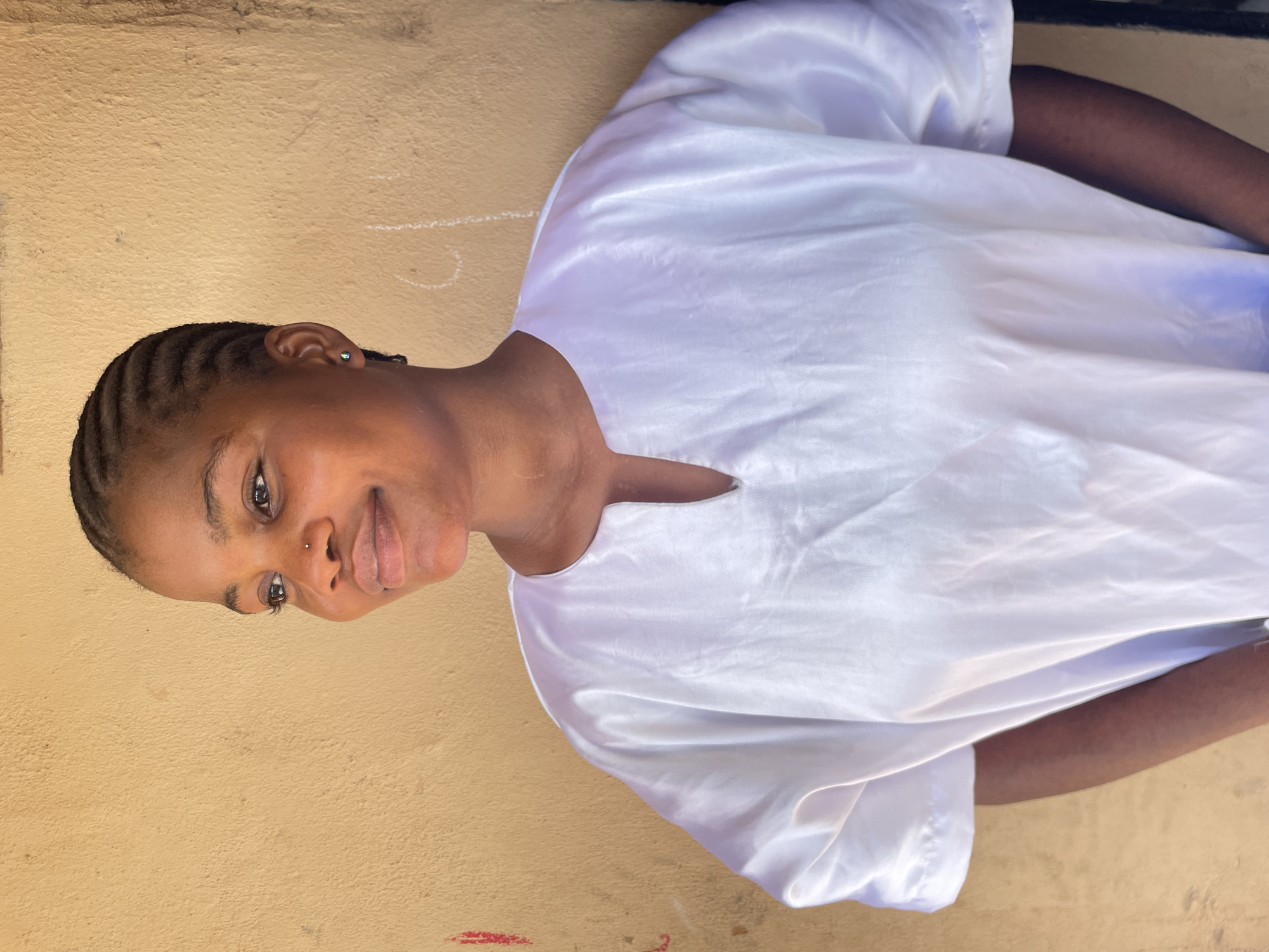 The image displays a portrait-style shot of a young individual, rotated 90 degrees clockwise. When viewed upright, it features a person with dark skin, framed from the chest up, positioned slightly to the left against a textured, light brown wall.
**Subjects:** The central subject is a young person with dark skin. Their hair is styled in neat cornrows or braids. They have a small, metallic stud piercing in their right nostril and a small, round earring in their left earlobe. The individual is wearing a loose-fitting, white, shiny garment, likely made of satin or a similar fabric, featuring a simple V-neckline.
**Activity or event taking place:** The person is posing for a photograph. They are looking slightly upwards and to their right, with a gentle, subtle smile.
**Notable details:**
* **Emotions:** The person exhibits a calm and pleasant expression, with a soft smile and bright, engaging eyes.
* **Background:** The background consists of a rough, textured wall, possibly finished with plaster or concrete, painted in a light brown or tan hue.
* **Wall markings:** Faint white chalk-like scribbles and simple shapes (such as a "U" and a dotted line) are visible on the wall, particularly in the upper and left sections (when viewed upright). A small red smudge or mark is also present on the wall, towards the bottom left.
* **Lighting:** The lighting is bright and natural, suggesting daytime. The absence of harsh shadows indicates diffused light, possibly from an overcast sky or an indirect light source.
* **Setting:** The textured wall and informal markings suggest an outdoor or semi-outdoor setting. This casual backdrop is consistent with a location such as Badagry, Nigeria, providing an authentic context for the portrait.
* **Visible text:** There is no discernible text visible in the image.