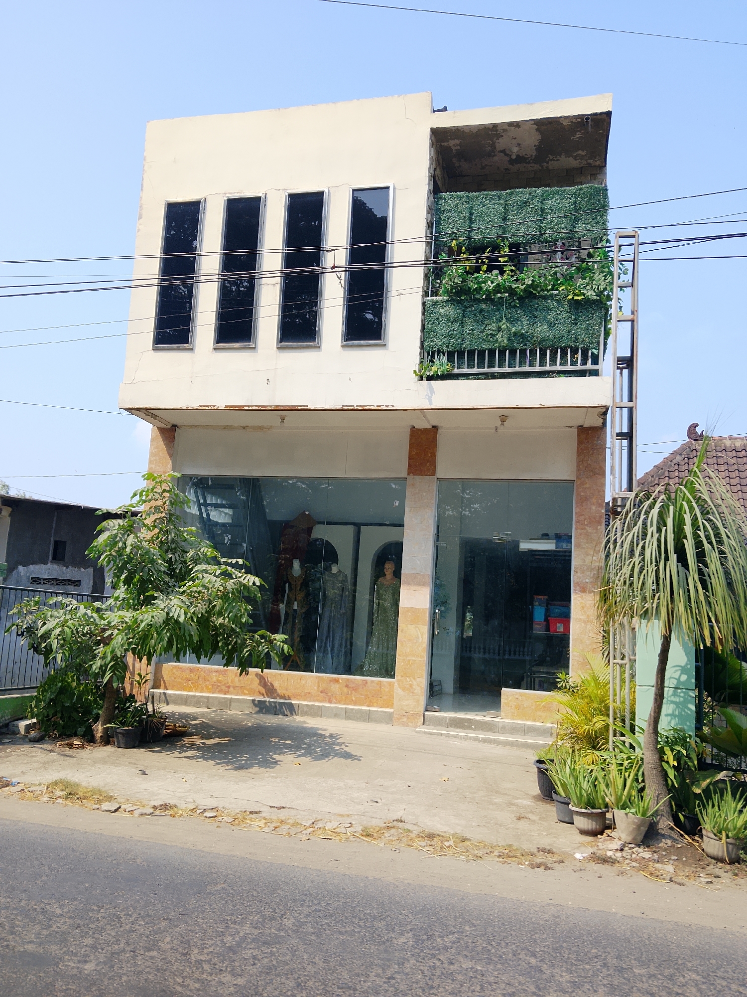The image shows a three-story building with a cream-colored facade, located in the city of Ngelo, Indonesia. The ground floor features a full glass storefront, allowing a view of mannequins dressed in traditional clothing inside, along with shelves of stacked boxes. The second floor has a similar glass facade, while the third floor is partially obscured by a balcony covered in green artificial foliage, with a metal ladder attached to the exterior. Several power lines cross the sky in front of the building. The building is situated next to a road, with potted plants, including aloe vera and palm trees, lining the sidewalk in front of the storefront. To the left of the building, a smaller, weathered structure is visible. The sky is clear and blue, suggesting it is daytime.
