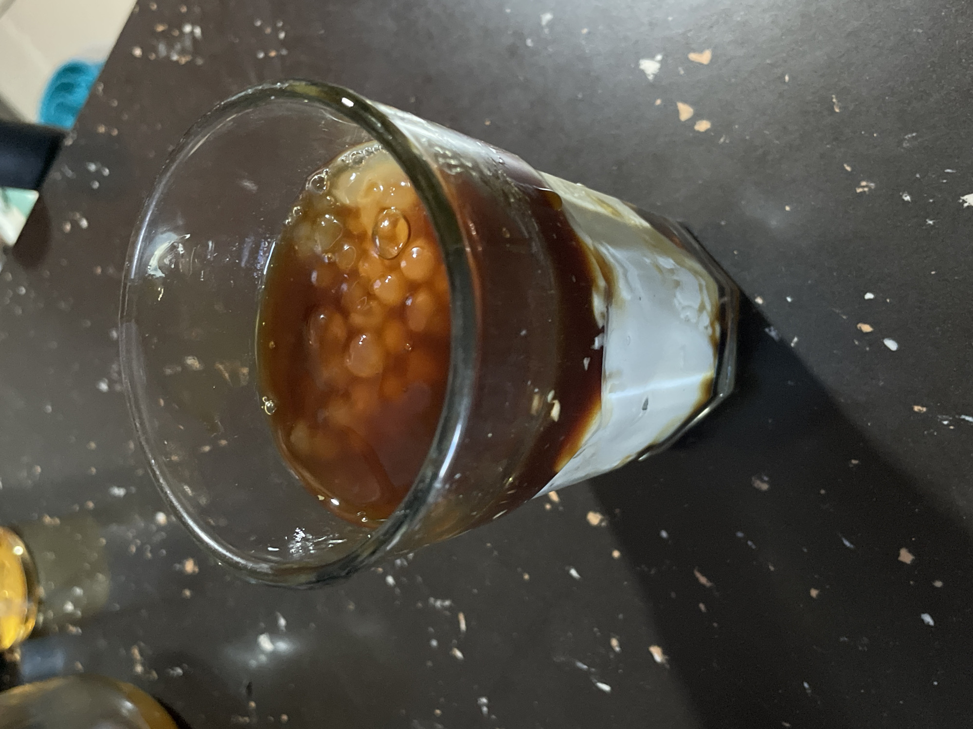 The image shows a glass of iced coffee with milk and tapioca pearls, likely a bubble tea or similar beverage. The drink is layered, with a white liquid at the bottom, a dark brown liquid in the middle, and a topping of small, translucent spheres, which are the tapioca pearls, suspended in a light brown liquid. The glass is clear and has a slight sheen. The drink is placed on a dark, speckled surface, possibly a countertop, which has small white and brown flecks. In the background, out of focus, are glimpses of other objects, including what appears to be a yellow liquid in a clear container on the left. The lighting suggests an indoor setting, possibly a home kitchen or a cafe.
There is no visible indication of the city of Davao City, Philippines in the image.