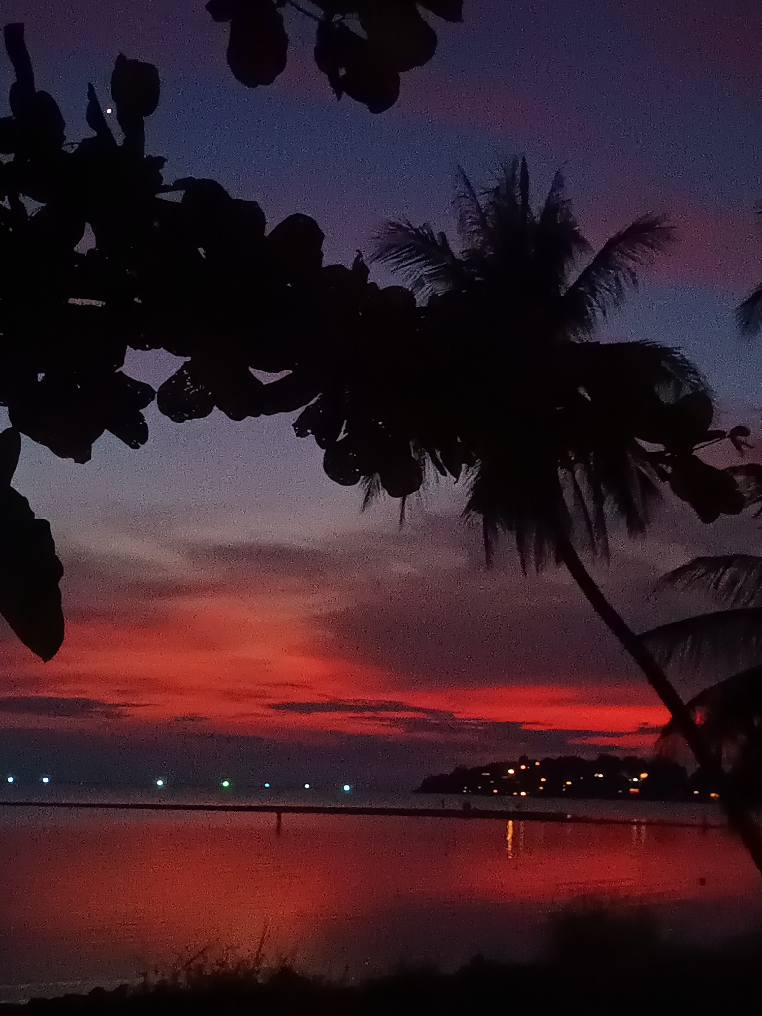 The image depicts a sunset scene in Baan Sri Thanu, Thailand. In the foreground, silhouettes of tree branches and palm trees frame the view. The sky transitions from a deep blue at the top to a vibrant red and orange near the horizon. The water reflects the colors of the sky, creating a mirror-like effect. Across the water, a landmass is visible with scattered lights, suggesting a settlement. A series of evenly spaced lights appear along a pier or structure extending into the water. The overall scene conveys a tranquil, tropical evening atmosphere.