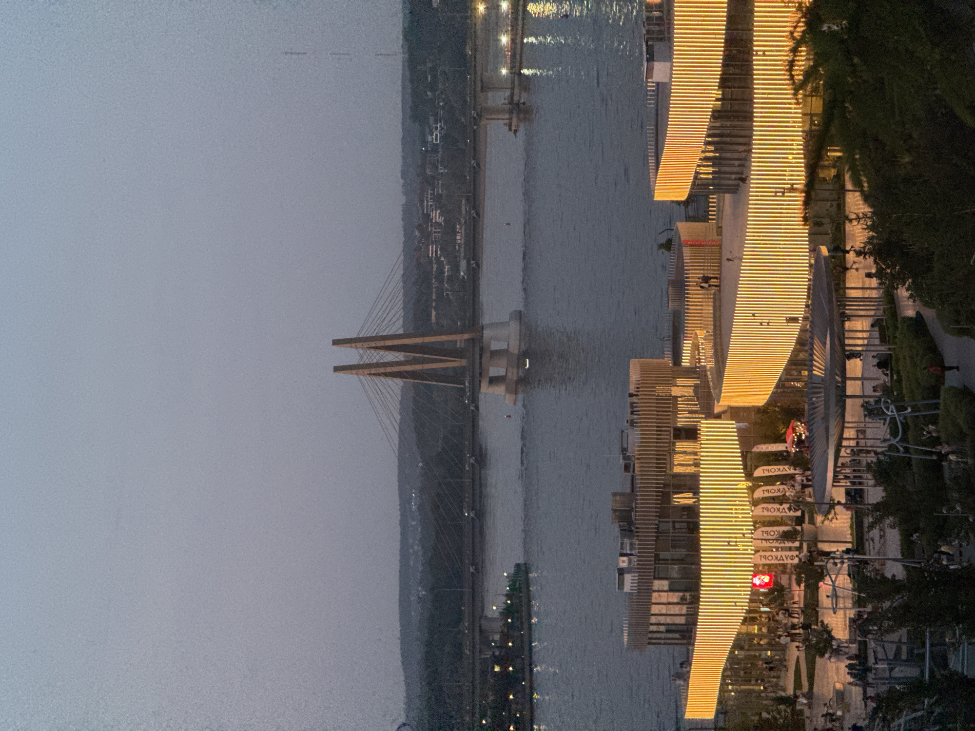 The image depicts a cityscape at dusk in Kazan, Russia. In the distance is the Millennium Bridge, a cable-stayed bridge spanning the Kazanka River. To the right of the bridge is a cluster of buildings with prominent horizontal illuminated lines, possibly part of the Kazan Riviera area. A waterfront park or promenade runs along the riverbank, with visible trees, pathways, and people. The sky is overcast, and the water surface reflects the city lights.