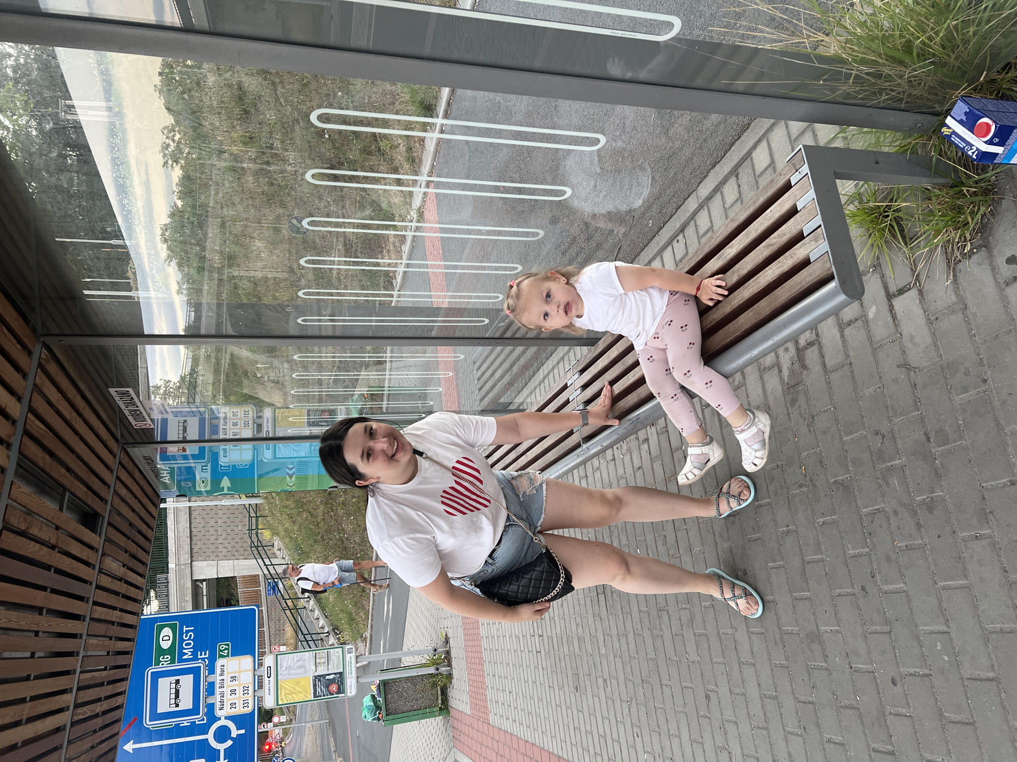 A young woman wearing a white t-shirt with a red heart graphic and denim shorts is bending down, smiling and looking at the camera. She has a black quilted purse with a gold chain strap. Next to her, a young girl with blonde pigtails is sitting on a wooden bench, wearing a white t-shirt and pink patterned leggings. The scene appears to be a bus stop in Pilsen, Czechia, evidenced by the signage with Czech text and route numbers such as "49". The bus stop features a glass enclosure with white loop designs and a wooden slatted bench. In the background, a man with a backpack is walking up a grassy slope next to a staircase. A blue and white carton of juice is visible on the ground next to the bench.