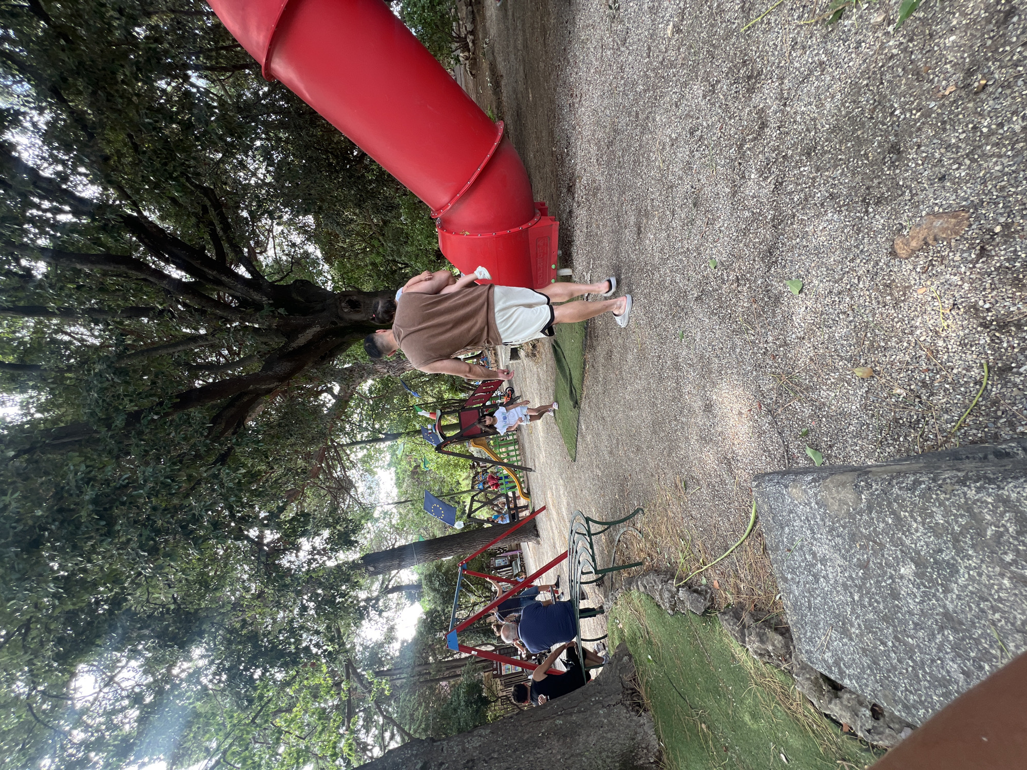 The image depicts a playground in Palmi, Italy, featuring a large red slide. In the foreground, a man in a brown sleeveless shirt and white shorts carries a young child. The playground is equipped with swings and other play structures, visible in the background. Several people are scattered throughout the scene, some sitting on benches and others near the play equipment. The environment is characterized by trees and a gravelly ground surface.