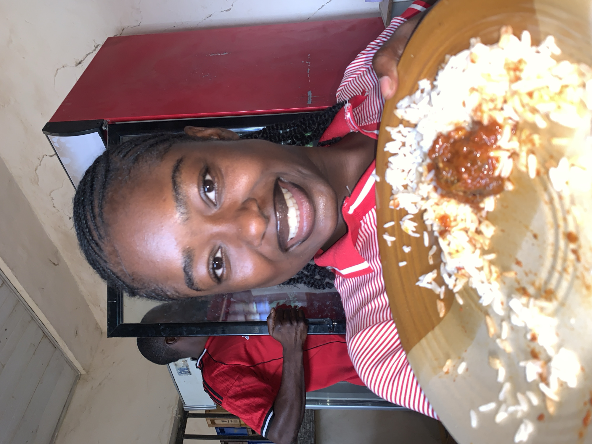 This media file captures an indoor scene, likely a casual eatery or canteen, in the vicinity of New Stadium, Nigeria. The primary subject is a person with dark skin and braided hair, wearing a red and white striped collared shirt, who is smiling broadly at the camera. They are holding up a round, brown plate filled with white rice and a central portion of reddish-brown sauce or stew.
In the background, a large red refrigerator is visible. Below the main subject, a reflection in what appears to be a glass display counter or polished surface shows another person with dark skin, wearing a red short-sleeved shirt with black stripes. This individual is seen from behind, positioned behind the counter.
The main subject's wide smile conveys a sense of happiness or enjoyment, possibly related to the meal they are presenting. The walls in the background exhibit some cracks, suggesting a well-used space. The lighting is bright and even, indicating an indoor setting during the day or a well-lit interior. No discernible text is visible on any objects in the image.